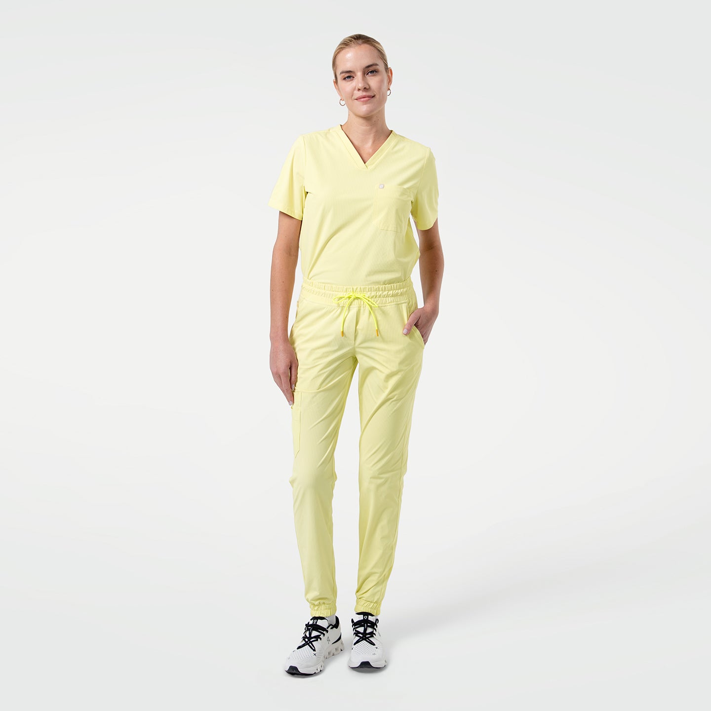 Image of Standing with one hand in the pocket, the model wears a Pineapple Yellow V-neck scrub top with short sleeves and a chest pocket, paired with matching jogger-style scrub pants and white sneakers against a clean light background.