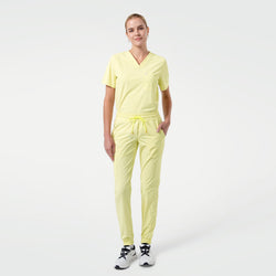 Image of Standing with one hand in the pocket, the model wears a Pineapple Yellow V-neck scrub top with short sleeves and a chest pocket, paired with matching jogger-style scrub pants and white sneakers against a clean light background.