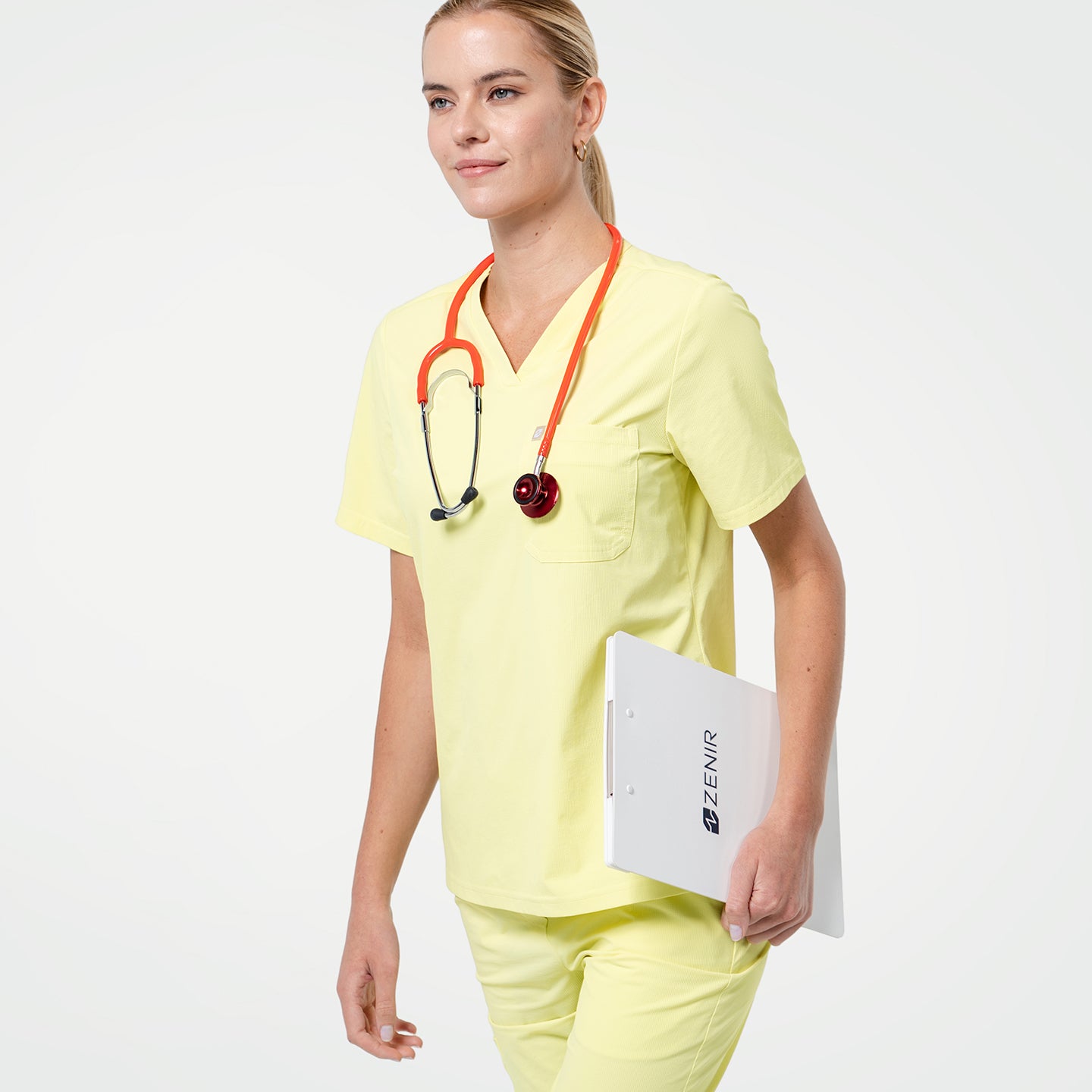 Walking pose in Pineapple Yellow scrub top with short sleeves and chest pocket, styled with matching scrub pants and a stethoscope around the neck.