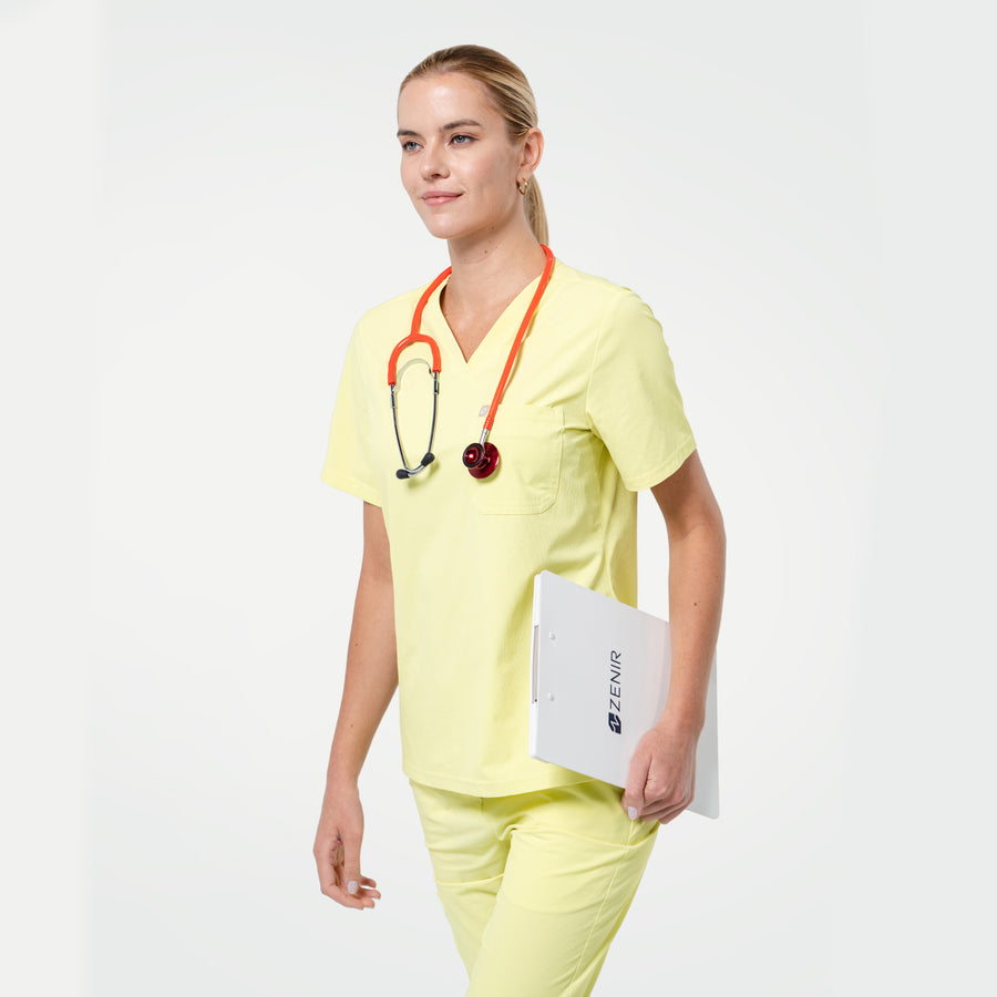 Walking pose in Pineapple Yellow scrub top with short sleeves and chest pocket, styled with matching scrub pants and a stethoscope around the neck.