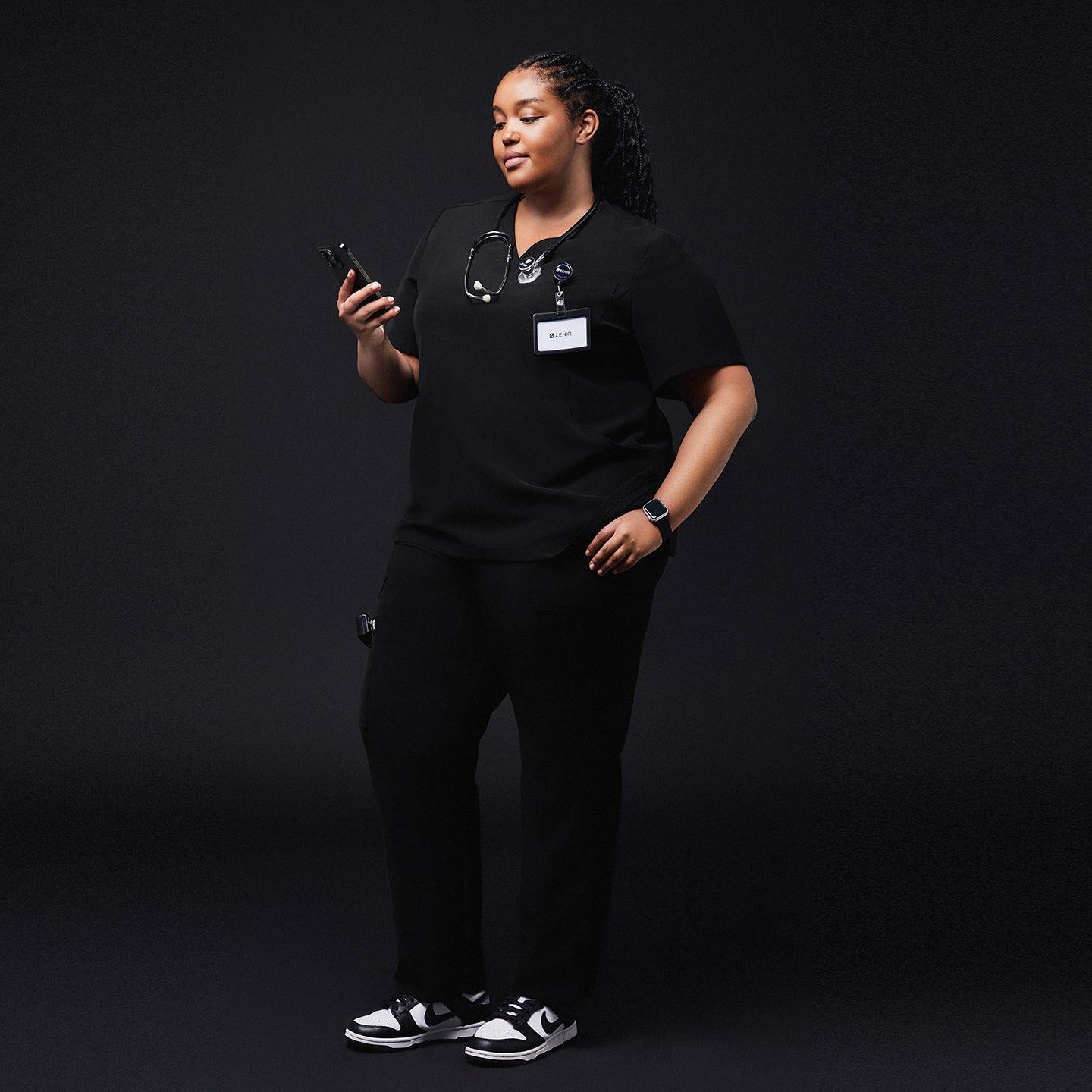 Image of Medical professional in black scrubs holding a smartphone, with a stethoscope around her neck and a name tag, standing confidently against a dark background,Rich Black