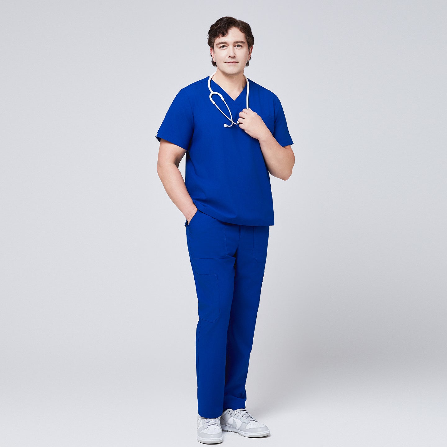 Image of Man wearing a royal blue V-neck scrub top with matching pants, holding a white stethoscope and standing in white sneakers,Royal Blue