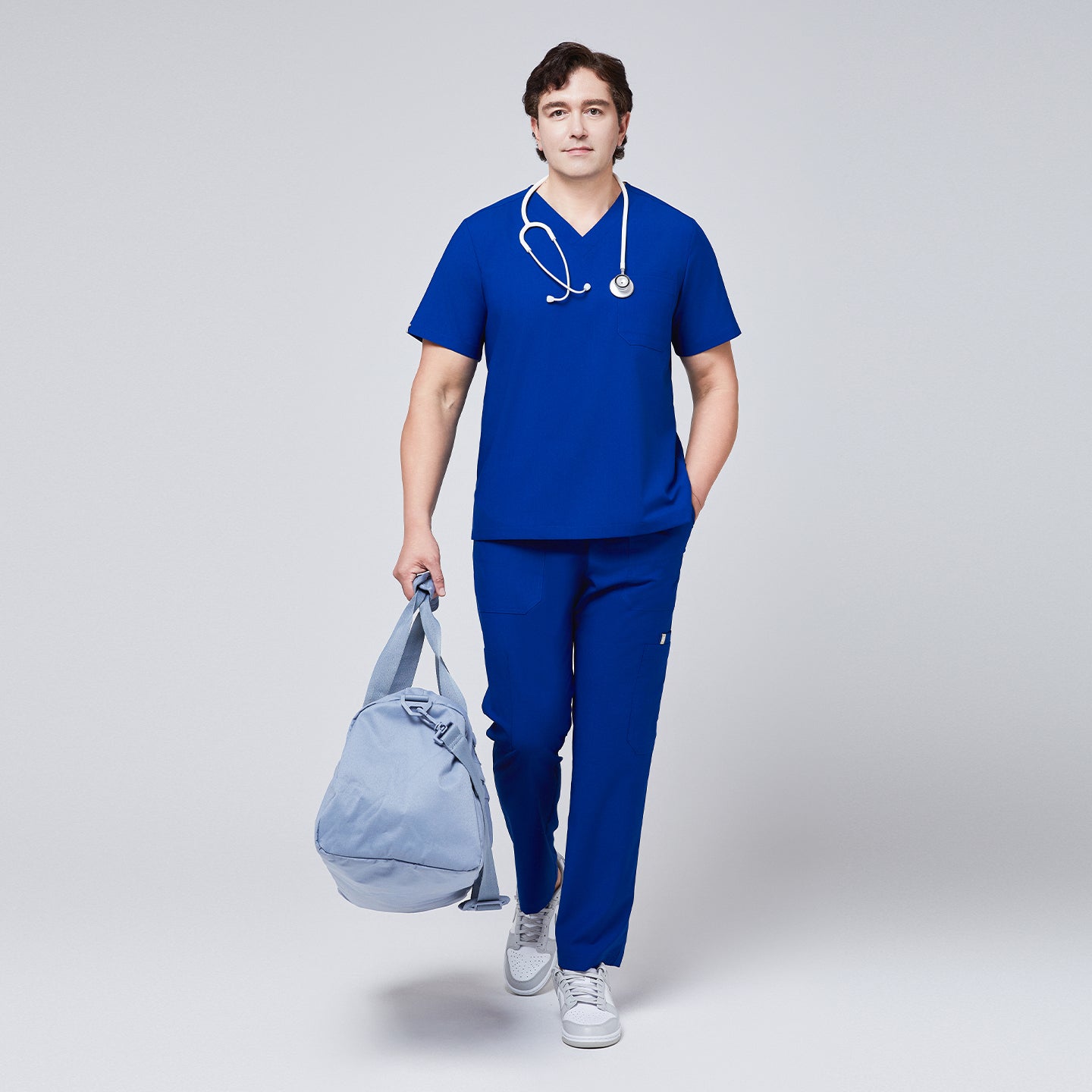 Man wearing royal blue V-neck scrub top and 8-pocket straight scrub pants, accessorized with a stethoscope and light blue duffle bag,Royal Blue