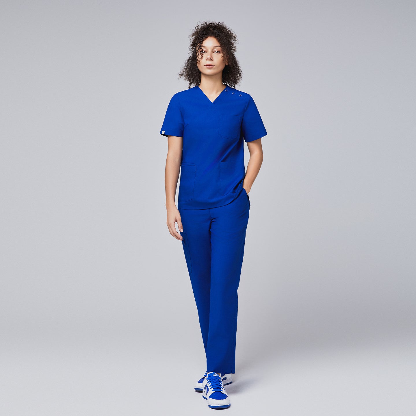 A woman dressed in a royal blue scrub set with button details on the shoulder, walking confidently with one hand in her pocket,Royal Blue
