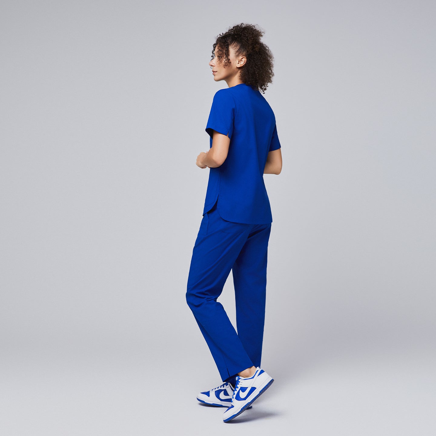 A woman modeling a royal blue scrub set with a relaxed pose, showcasing the back design of the V-neck top with shoulder button details and straight-leg pants,Royal Blue