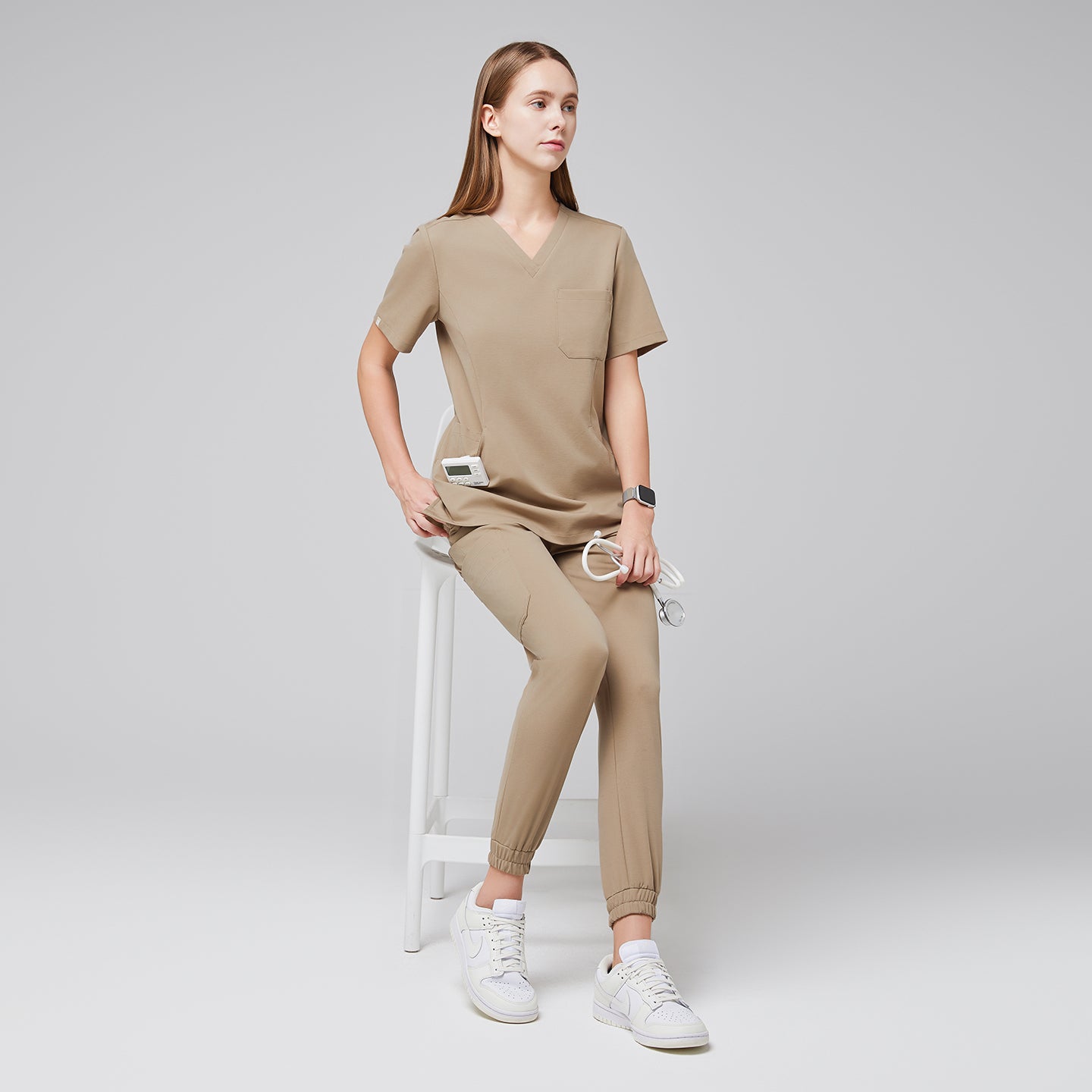 Image of Female wearing sand beige scrub top and jogger pants, sitting on a white stool, holding a stethoscope, paired with white sneakers,Sand Beige