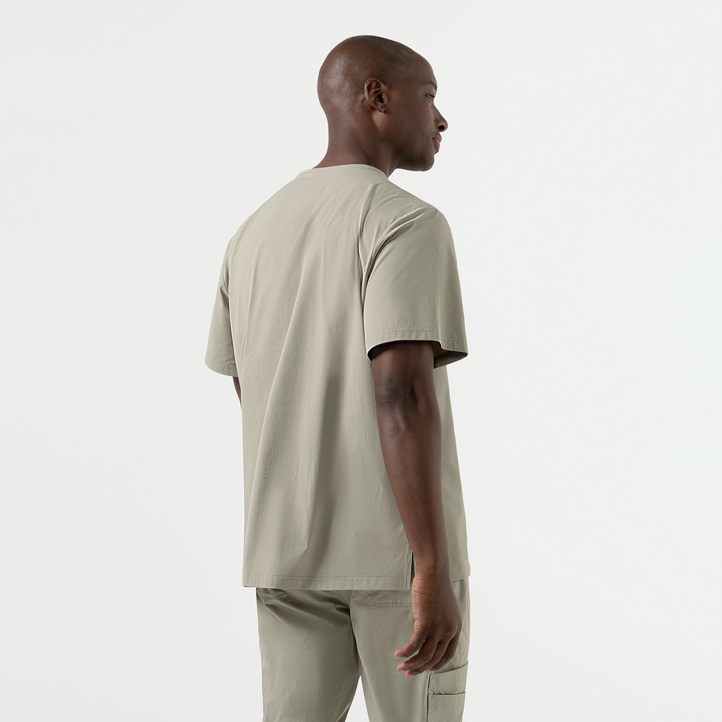 Back and side-angle view of a man wearing a Sand-Khaki single-pocket scrub top, highlighting the smooth, lightweight fabric, relaxed short-sleeve fit, and clean, minimal design that pairs seamlessly with matching scrub pants.