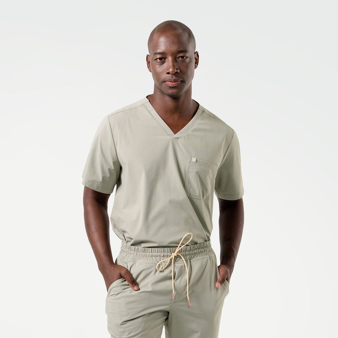 The male model is shown in Sand Khaki scrubs, standing with hands in pockets and facing forward, highlighting the V-neckline and clean lines of the top paired with drawstring jogger-style pants.