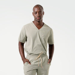 Image of The male model is shown in Sand Khaki scrubs, standing with hands in pockets and facing forward, highlighting the V-neckline and clean lines of the top paired with drawstring jogger-style pants.