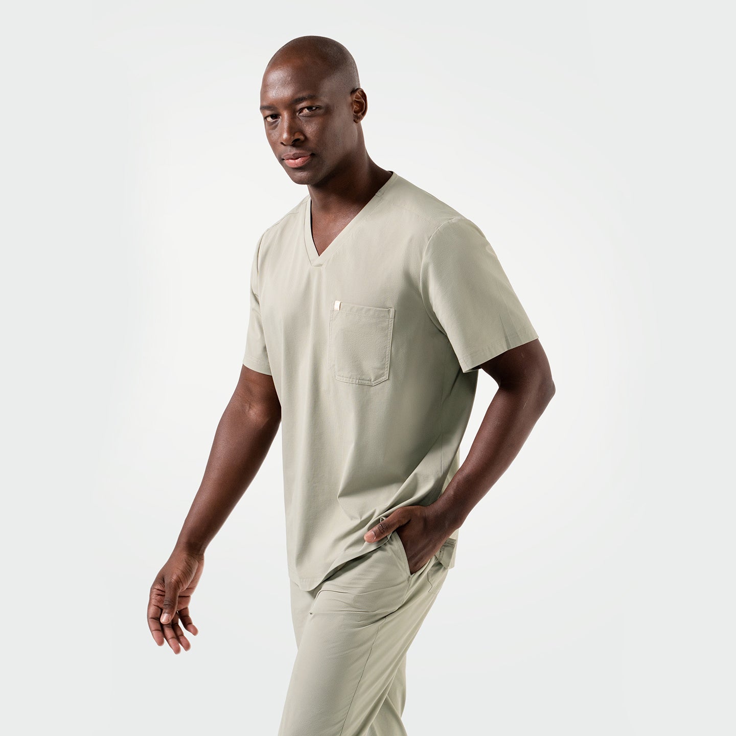 Image of Wearing the Sand Khaki Single Pocket Scrub Top, the model walks confidently with a composed look, hand in the pocket and body slightly angled.