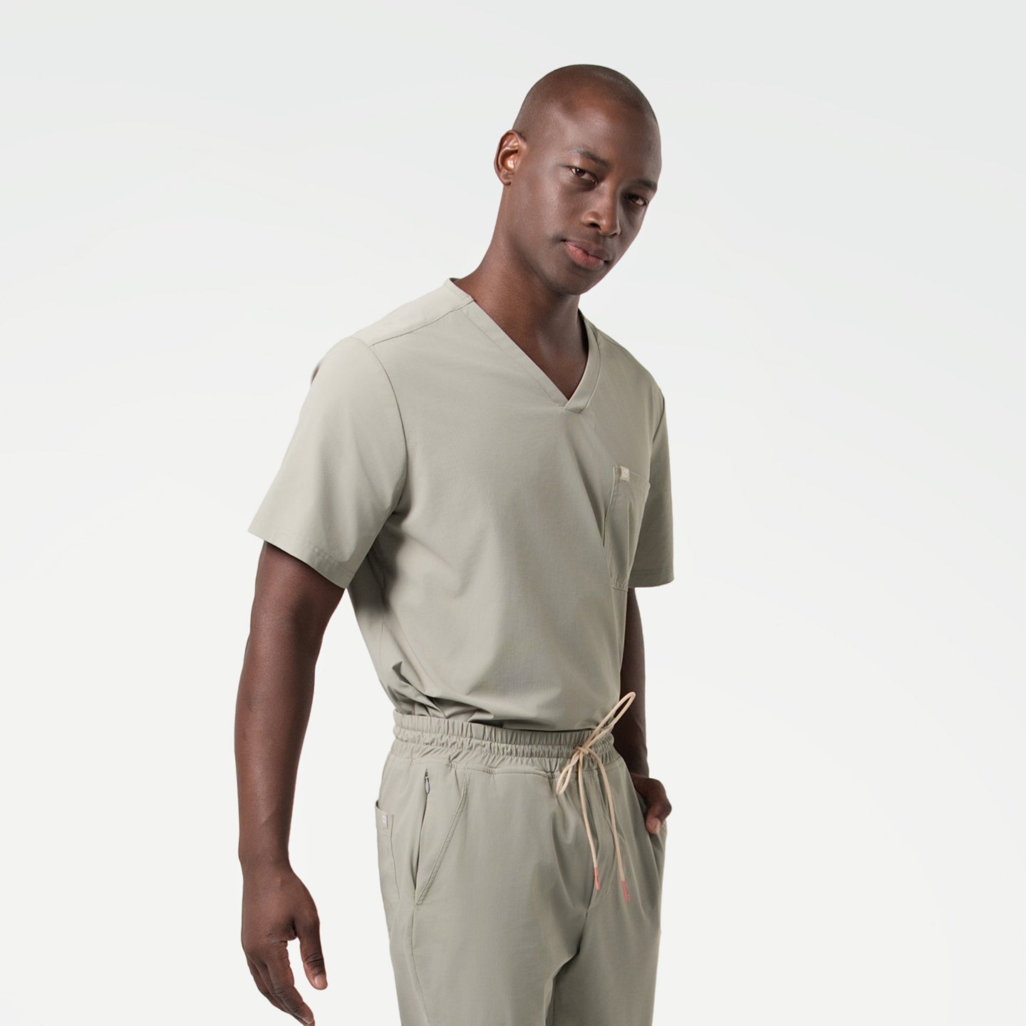 Wearing a Sand Khaki scrub set, the male model stands with a relaxed posture and one hand in his pocket, presenting a calm and professional look.
