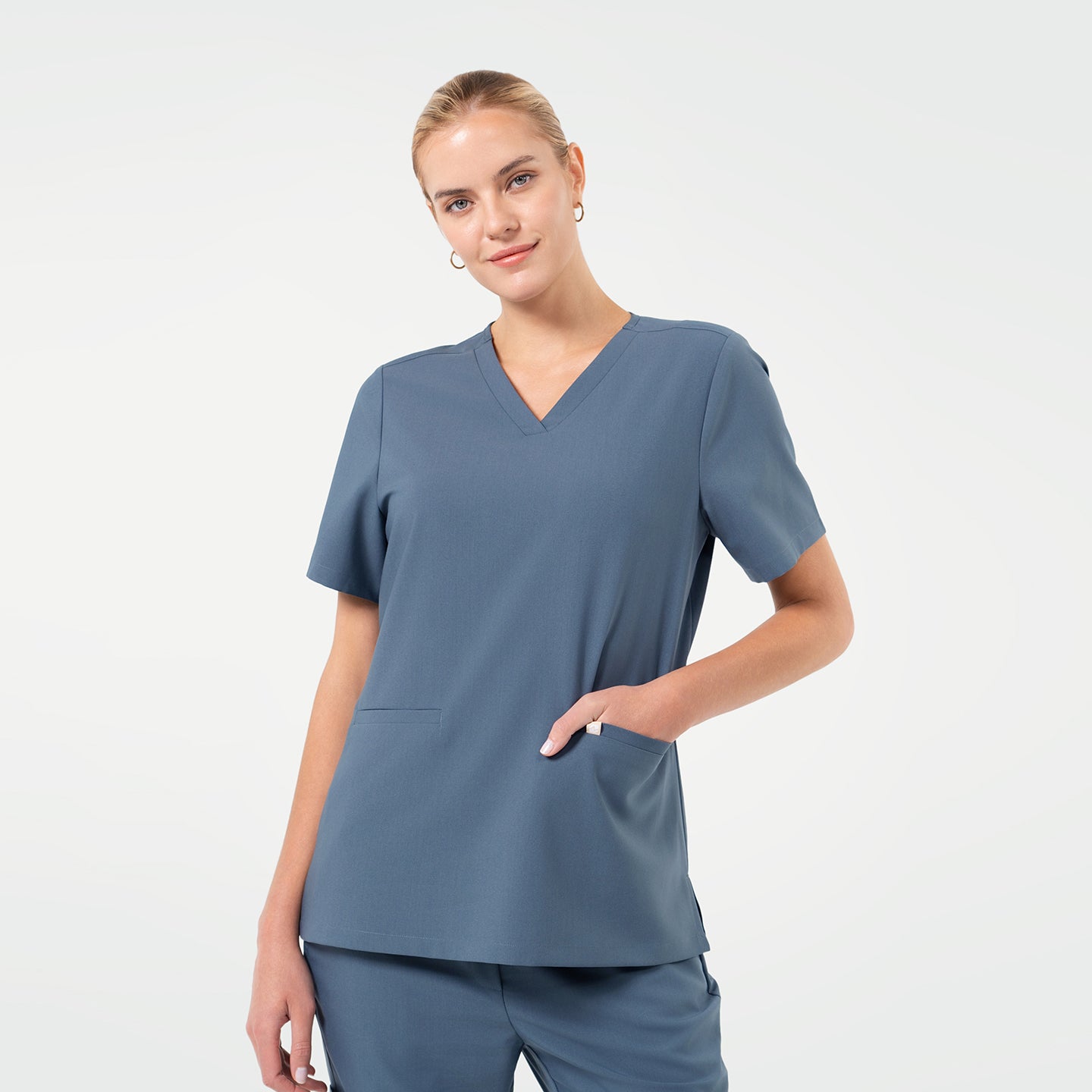 A female model poses with a soft smile while resting one hand in the pocket of a Smoky Blue V-neck scrub top, featuring short sleeves and a straight hem, paired with matching scrub pants against a clean light background.