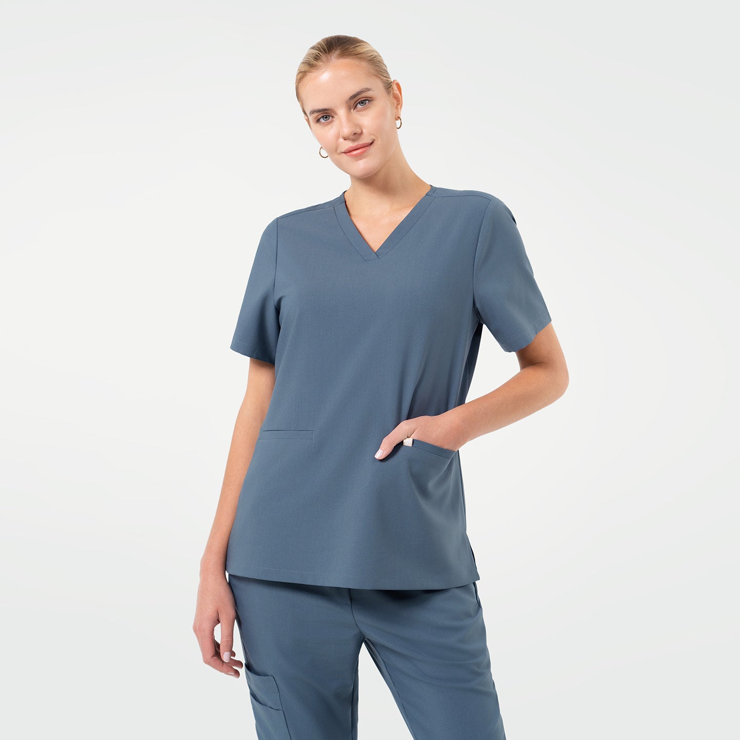 Image of A female model poses with a soft smile while resting one hand in the pocket of a Smoky Blue V-neck scrub top, featuring short sleeves and a straight hem, paired with matching scrub pants against a clean light background.