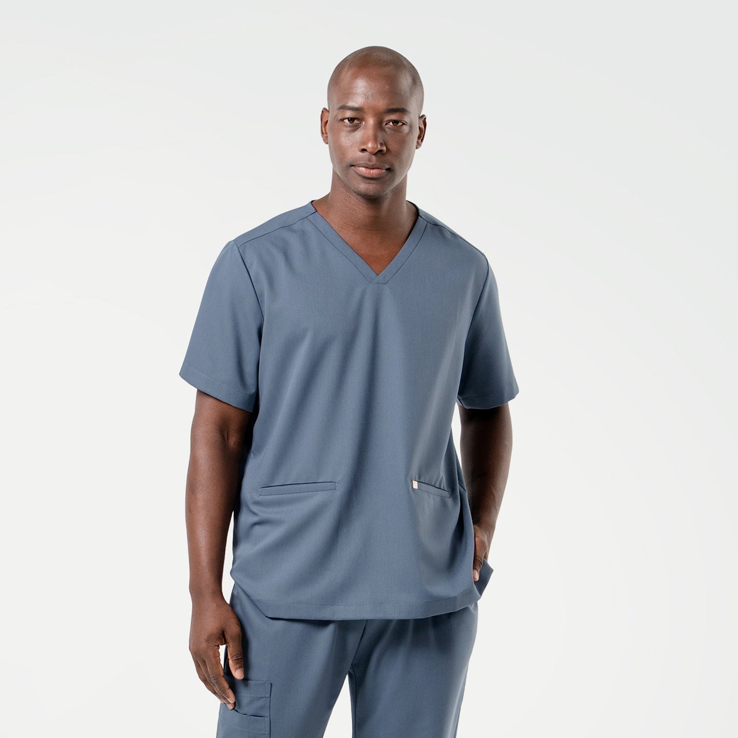 Wearing a Smoky Blue scrub set, the male model stands facing forward with one hand in his pocket, presenting a composed and professional look.