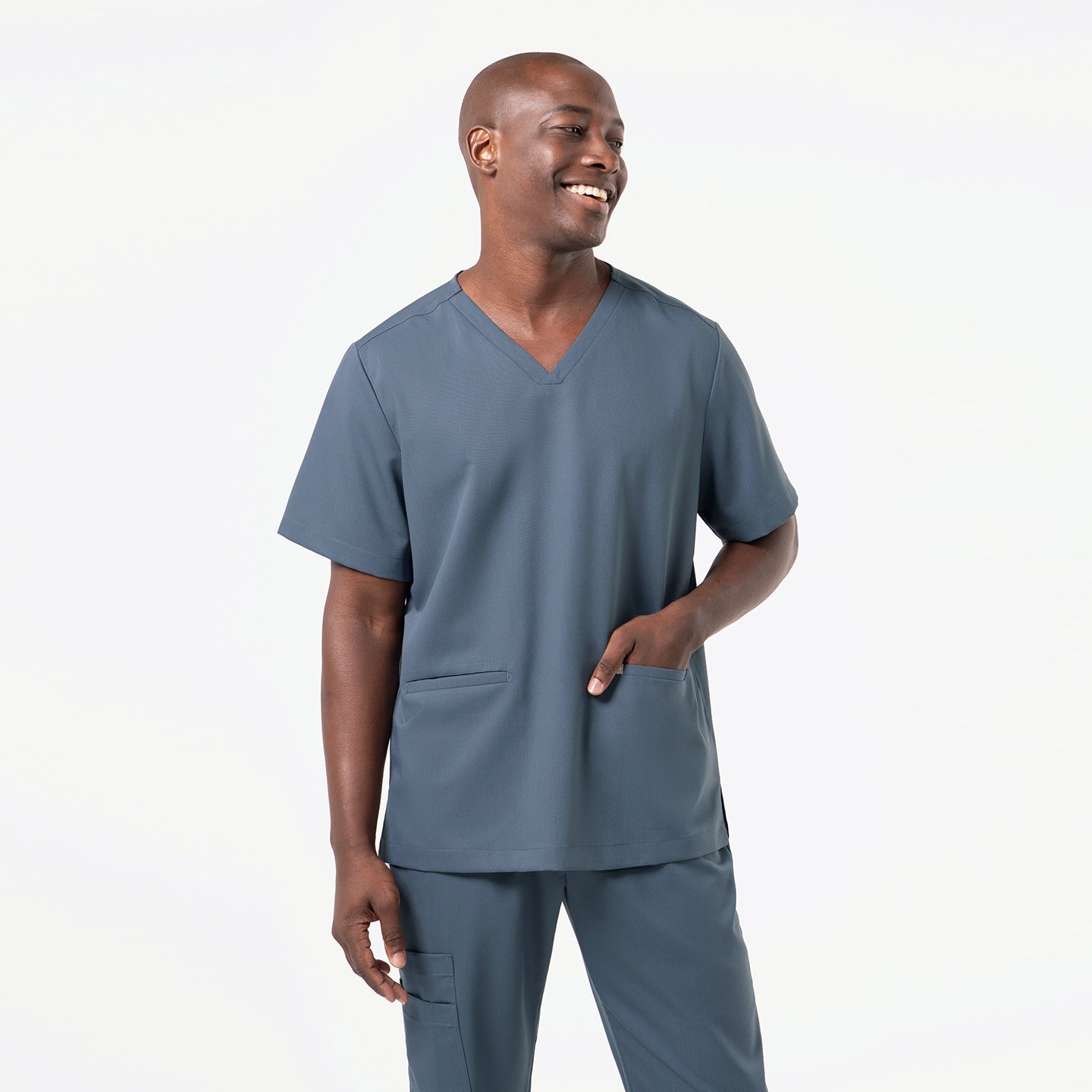 Smiling and standing in a relaxed pose, the model wears a Smoky-Blue double-pocket scrub top featuring a clean V-neckline, short sleeves, and two streamlined front pockets, highlighting the smooth, flexible fabric and comfortable, modern fit.