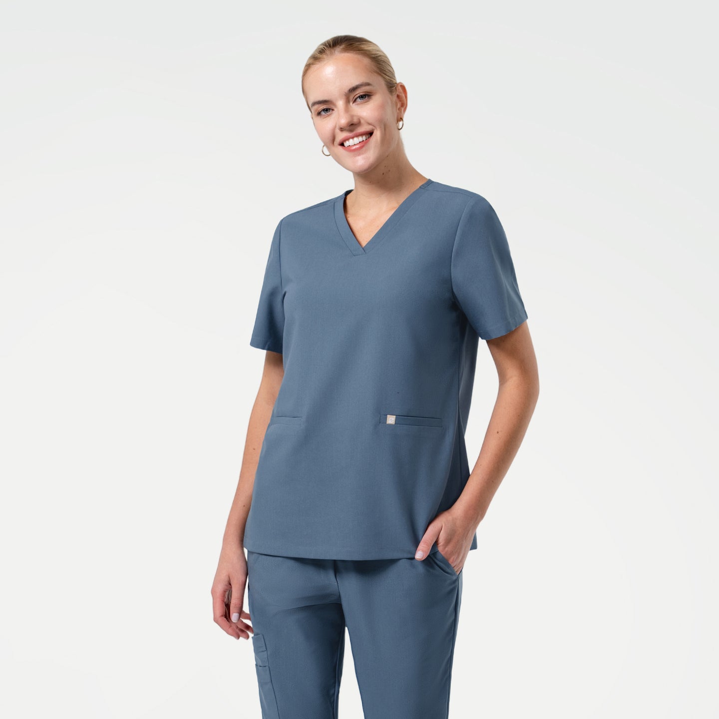 Image of In the Women's Smoky Blue Double Pocket Scrub Top, standing with a gentle smile, upper body shot