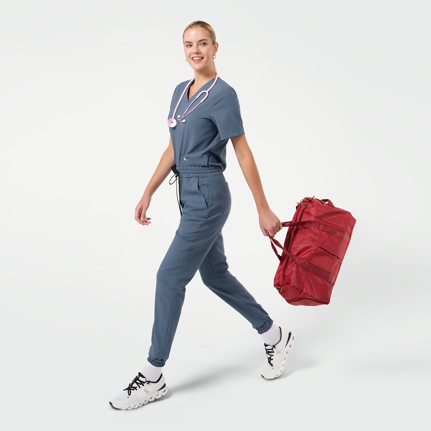 Image of A smiling female model walks forward carrying a red duffel bag while wearing Smoky Blue scrubs with a pink stethoscope around her neck and white sneakers.