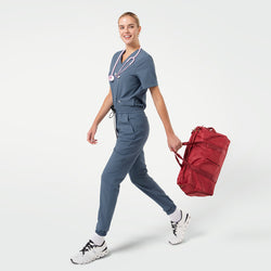 Image of A smiling female model walks forward carrying a red duffel bag while wearing Smoky Blue scrubs with a pink stethoscope around her neck and white sneakers.