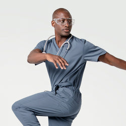 Image of In a Smoky Blue scrub set, the male model wears safety glasses and a stethoscope while extending his arms in a dynamic, balanced pose.
