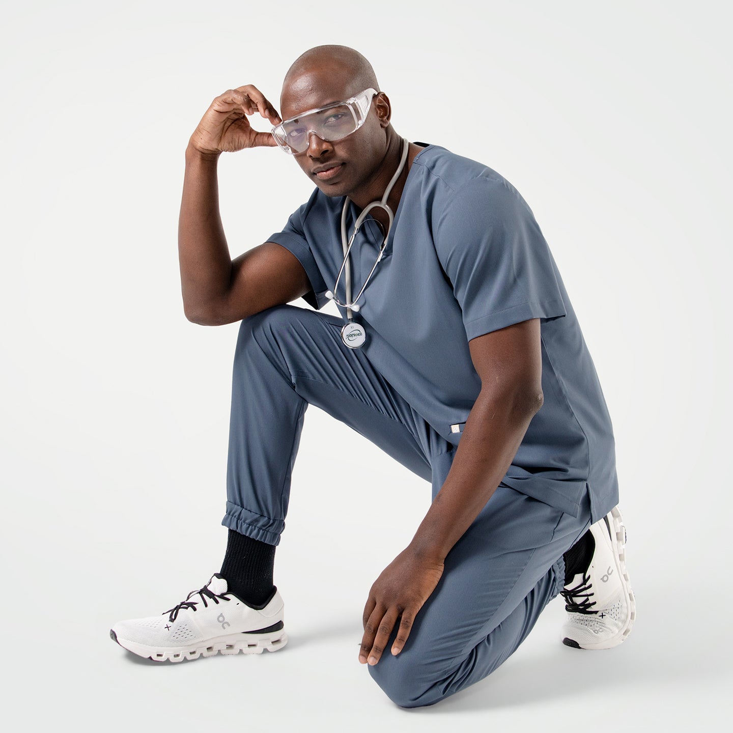 Dressed in a Smoky Blue scrub set, the male model kneels with one hand on his knee, wearing safety glasses and a stethoscope around his neck for a modern, professional look.