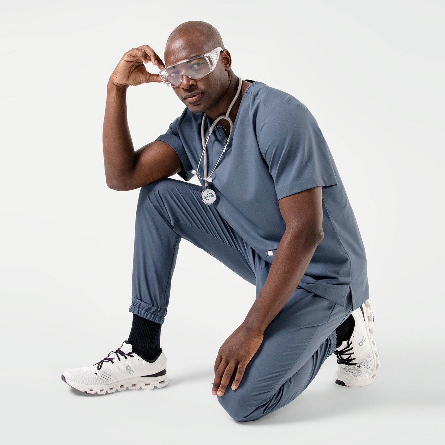 Image of Dressed in a Smoky Blue scrub set, the male model kneels with one hand on his knee, wearing safety glasses and a stethoscope around his neck for a modern, professional look.