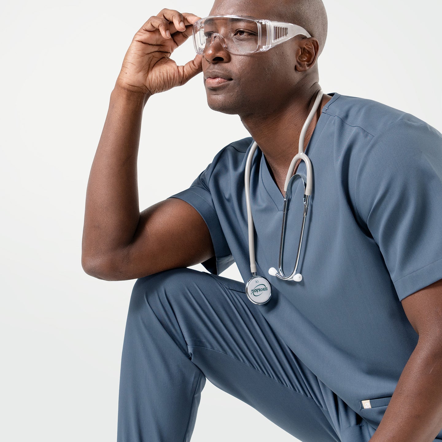 Image of In a Smoky Blue scrub set, the male model sits with one arm resting on his knee, wearing safety glasses and a stethoscope while gazing thoughtfully to the side.