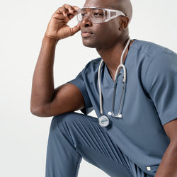 Image of In a Smoky Blue scrub set, the male model sits with one arm resting on his knee, wearing safety glasses and a stethoscope while gazing thoughtfully to the side.