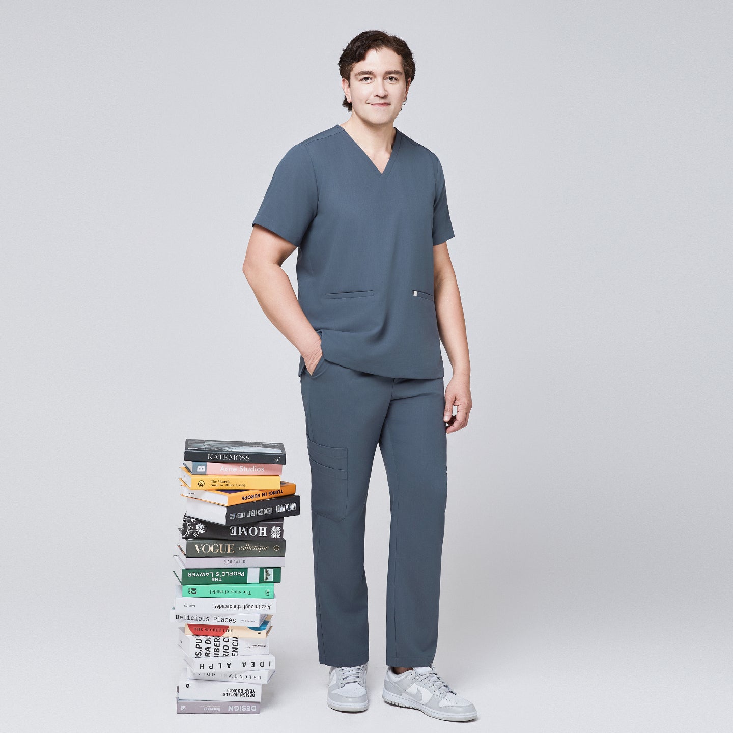 Image of Man in a smoky blue scrub set with a double-pocket V-neck top, standing next to a stack of colorful books,Smoky Blue