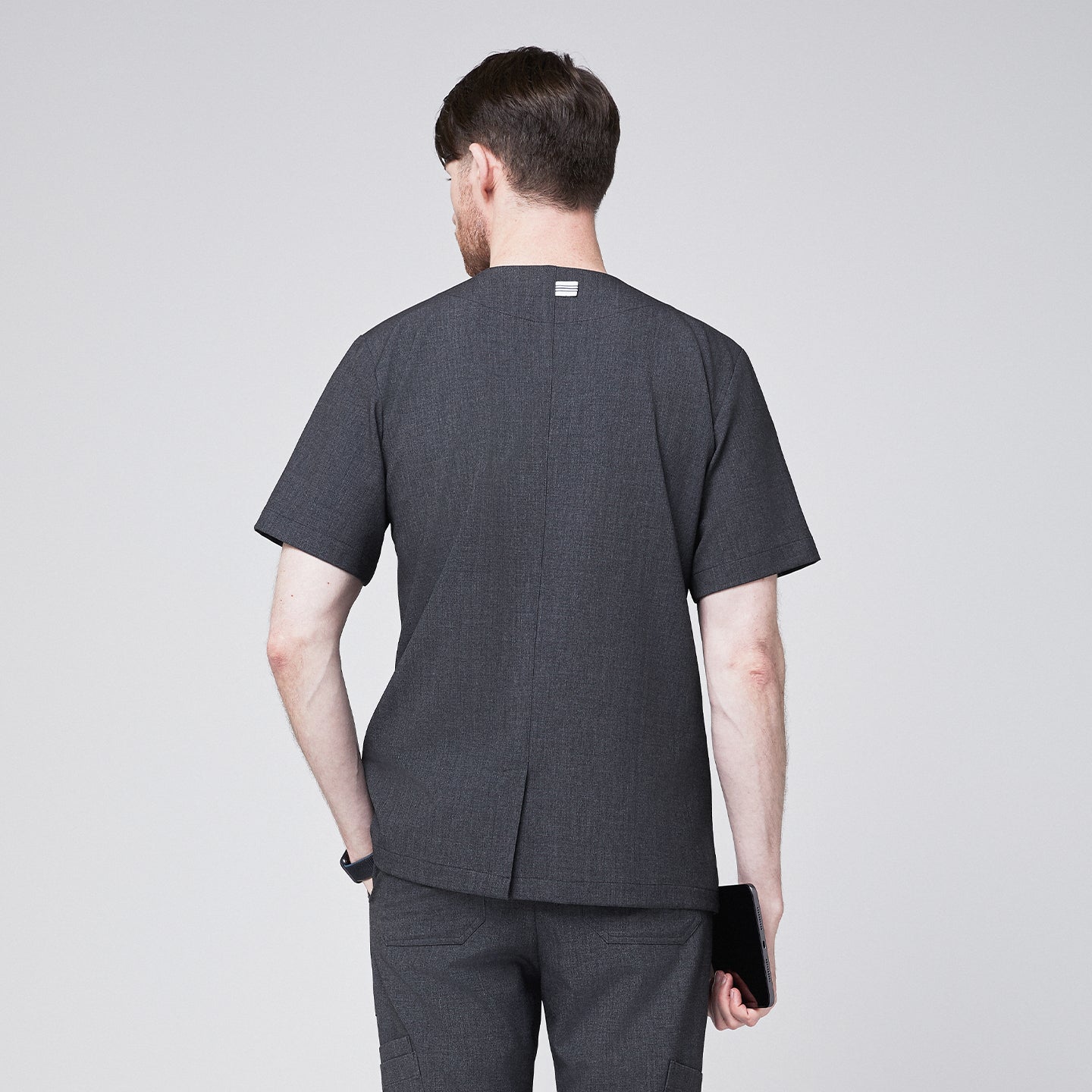 Man in a soft gray scrub top with a V-neck and shoulder button detail, viewed from the back, holding a tablet in one hand,Charcoal Gray