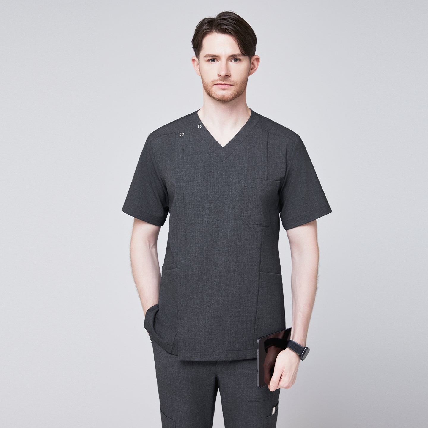 Image of Man wearing a soft gray scrub top with a V-neck and shoulder button detail, paired with matching pants, holding a tablet and posing with one hand in his pocket,Charcoal Gray