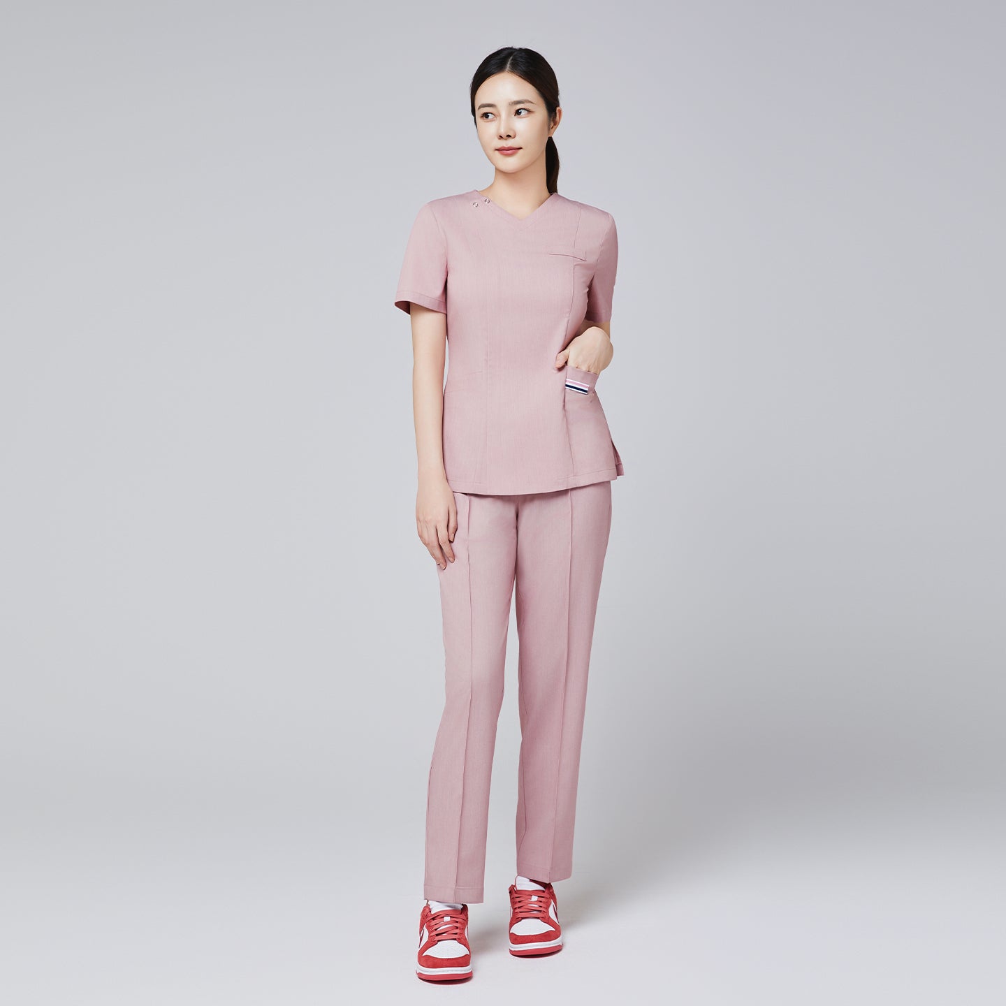 Image of Female model in a soft pink scrub set with front zipper detail, paired with red sneakers, posing with one hand in pocket,Soft Pink