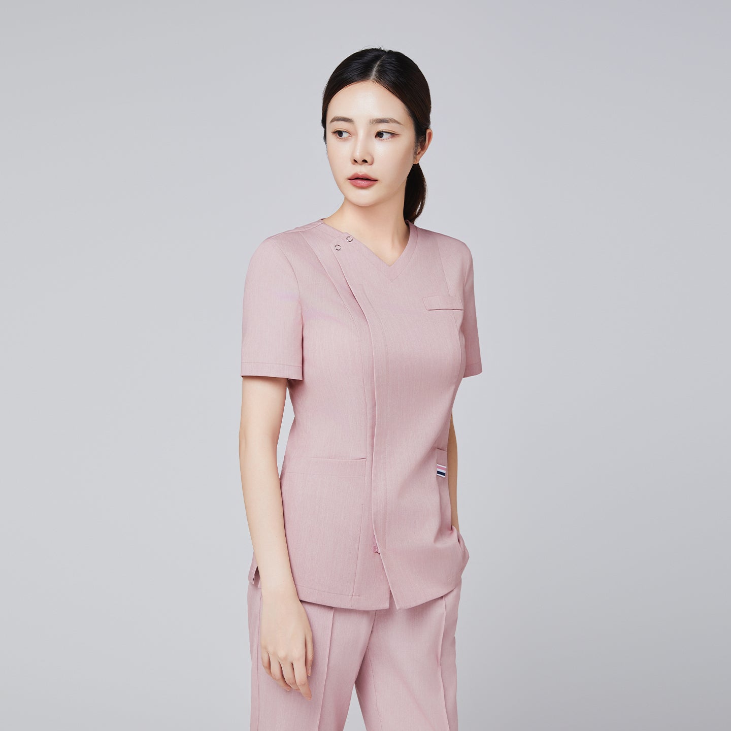 Female model in a soft pink scrub top with front zipper and tailored fit, paired with matching pants, looking to the side,Soft Pink