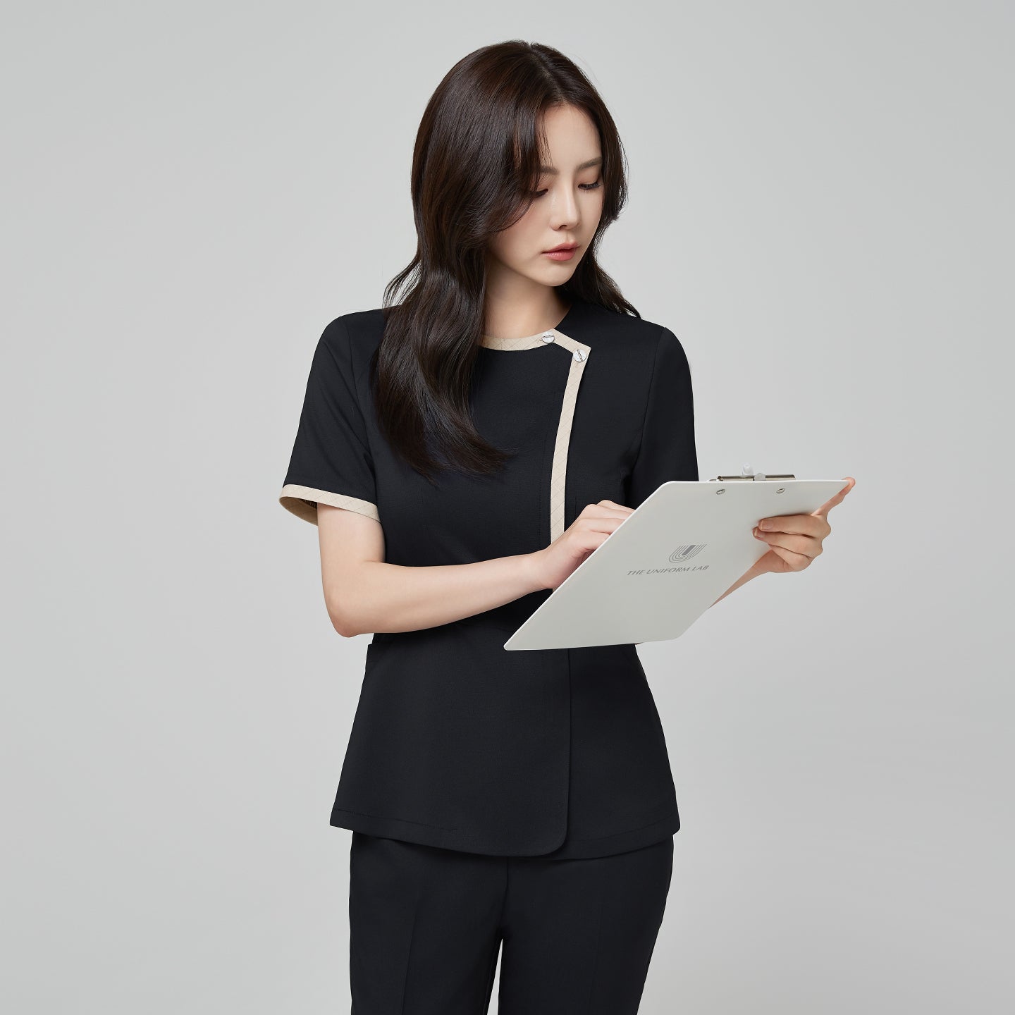 Model wearing TUL NS-175 Women’s Check Beige Round Collar Scrub Top in Stretch Black, showing contrasting beige trim along the neckline and front.
