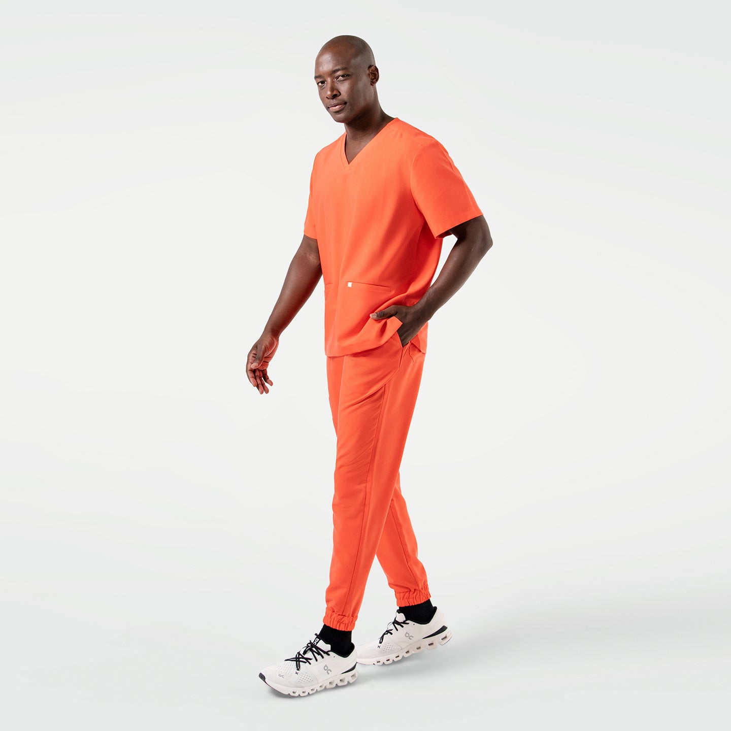 Image of Wearing the Tiger Orange Double Pocket Scrub Top, the model steps forward with one hand in the pocket.