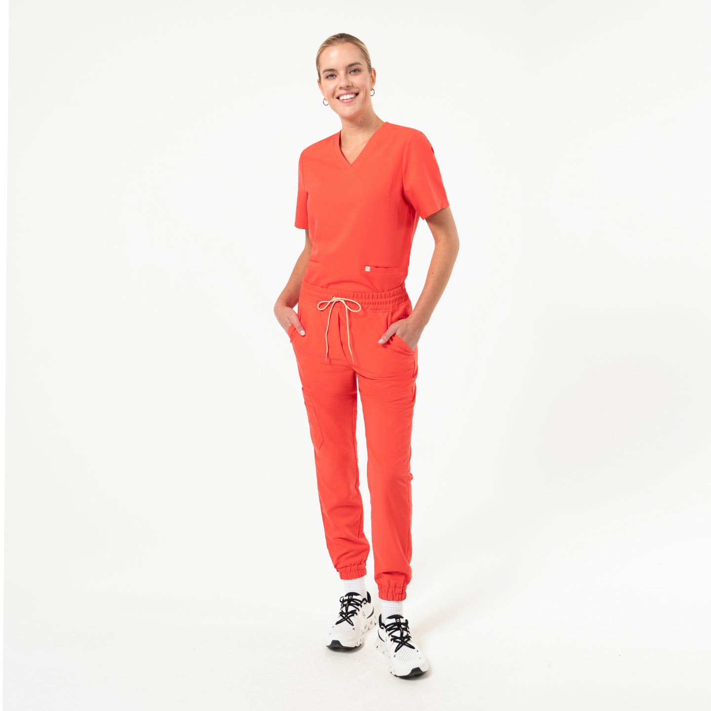 Model wearing a vibrant tiger-orange double-pocket scrub top and matching jogger pants, photographed in a minimal white studio background.