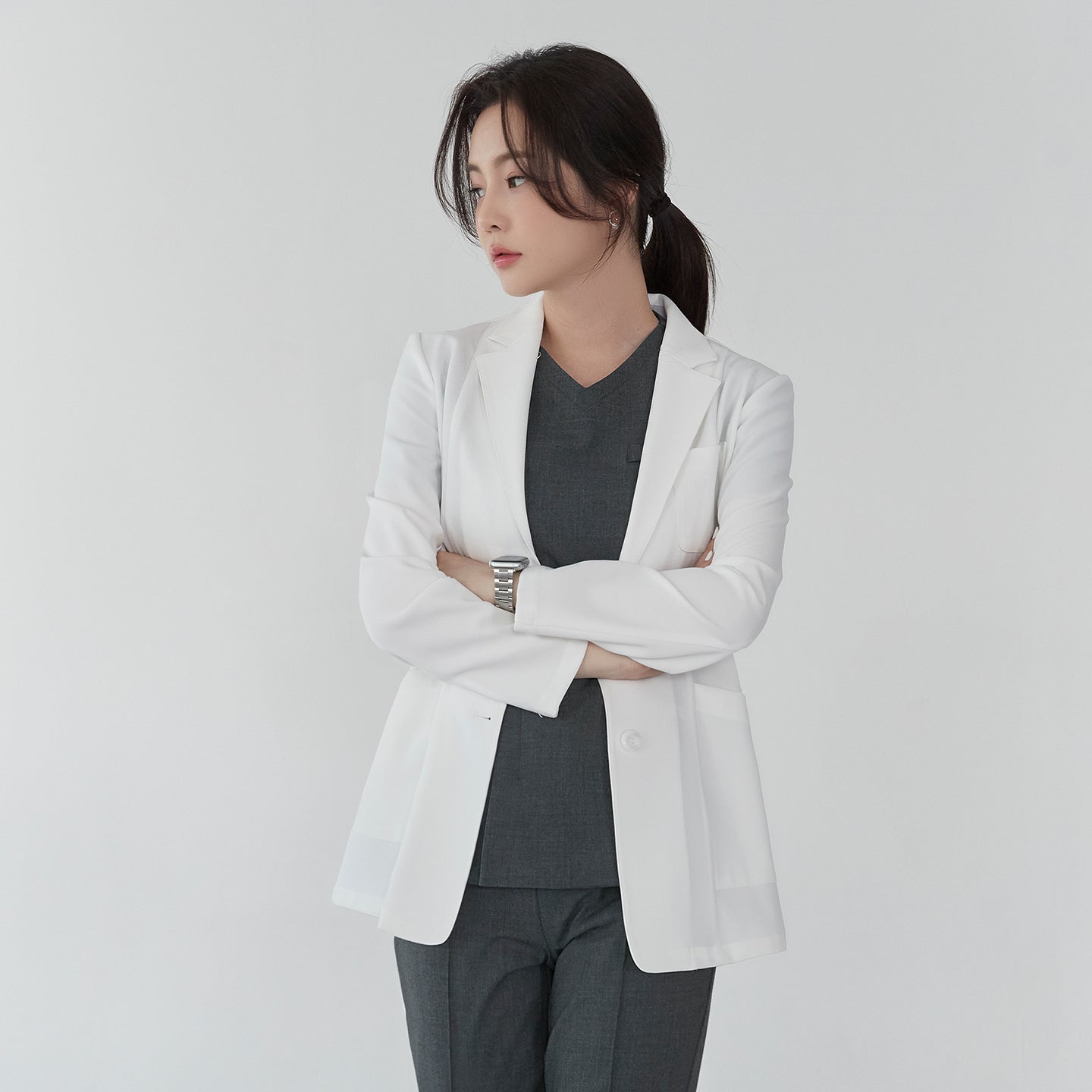 Front view of TUL LCW-01 White Ivory lab coat, worn by a model with arms crossed over a gray scrub top. The coat’s clean lines and fitted silhouette are visible, along with a single breast pocket.