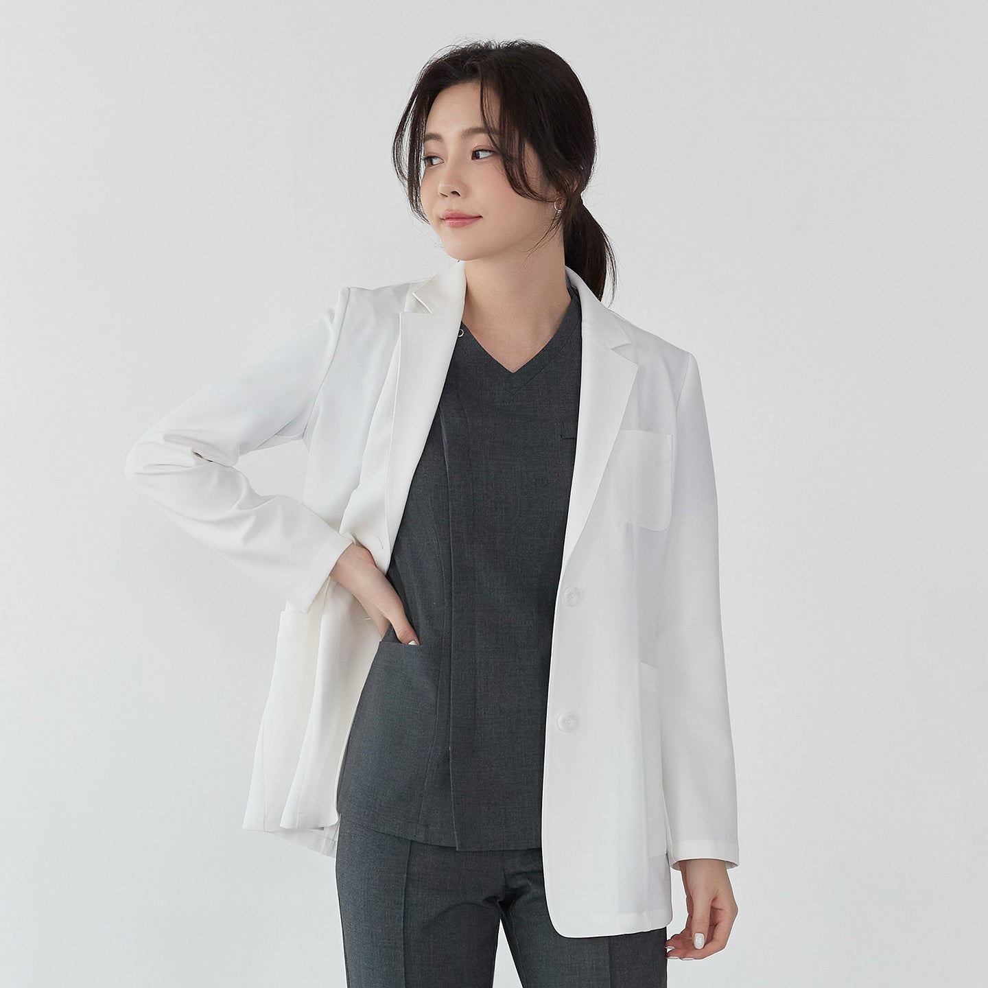 Model wearing TUL LCW-01 Women’s Short Lab Coat in White Ivory, casually posing with one hand in the coat pocket. The notched collar, button closure, and slightly curved hem are visible.