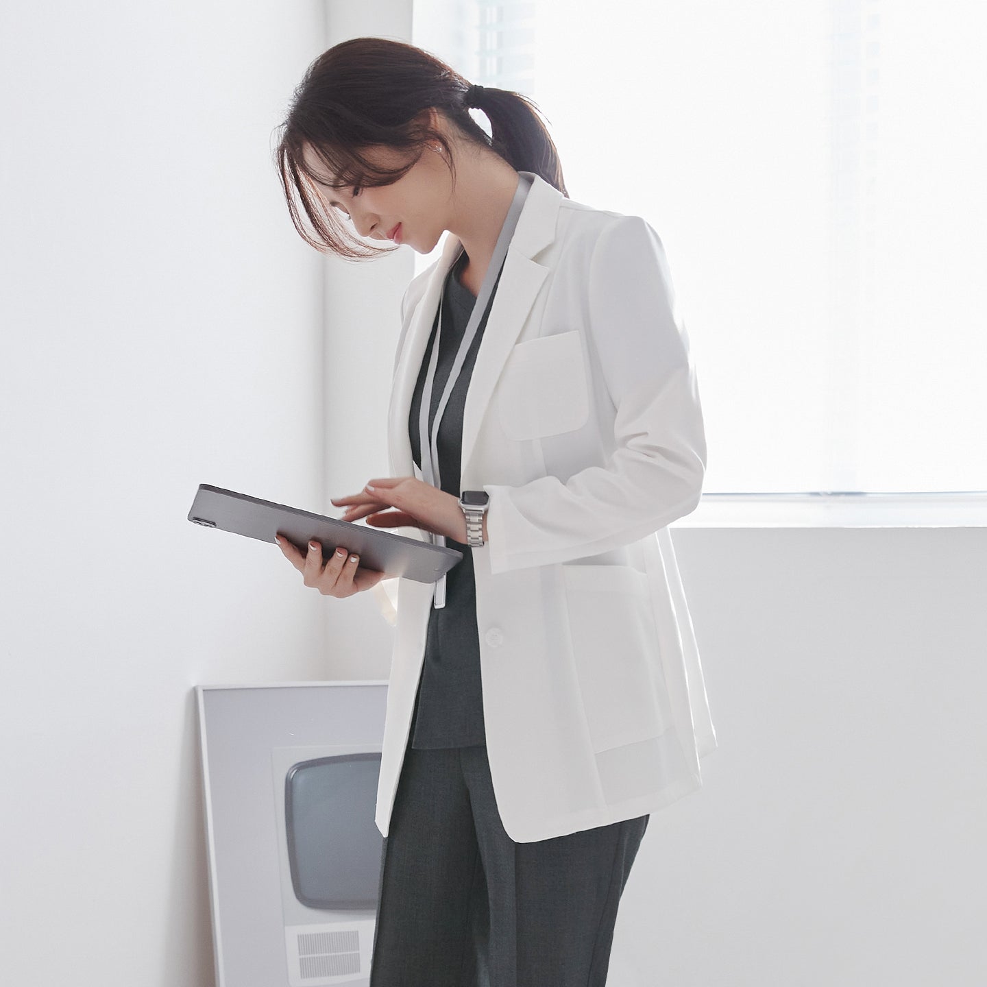 Model wearing TUL LCW-01 Women’s Short Lab Coat in White Ivory over gray scrubs, leaning forward to read from a tablet. The coat features a notched lapel, front pocket, and a slightly structured fit.