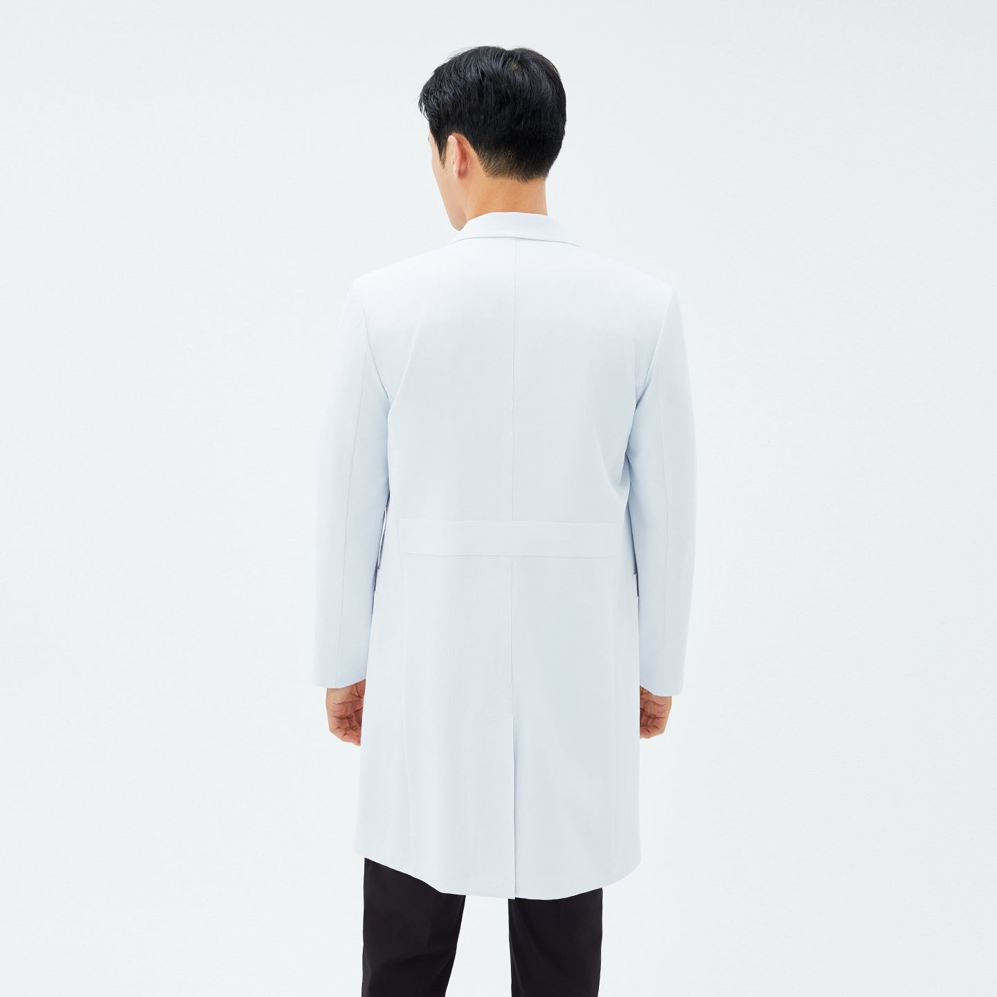 Back view of TUL LCM-11 Men’s Long Lab Coat in White, highlighting the structured waist seam and straight hem. The model’s black scrub pants are visible below the coat.