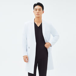 Image of Model wearing TUL LCM-11 White long lab coat over a black V-neck scrub top, posing with one hand in the coat pocket and the other adjusting the coat’s front.
