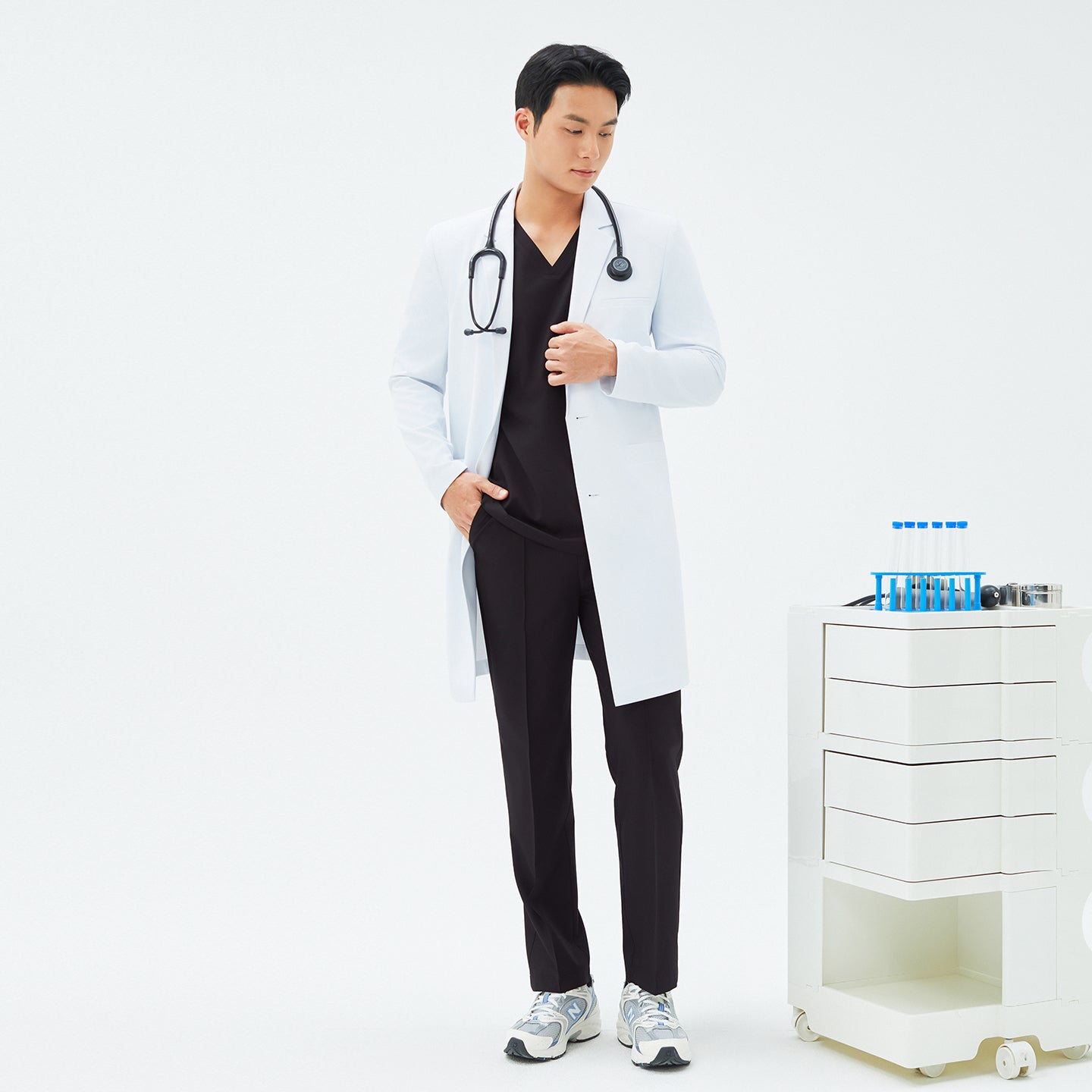 Full-length shot of a male model wearing TUL LCM-11 White long lab coat and matching black scrubs, standing next to a small white medical cart with test tubes.