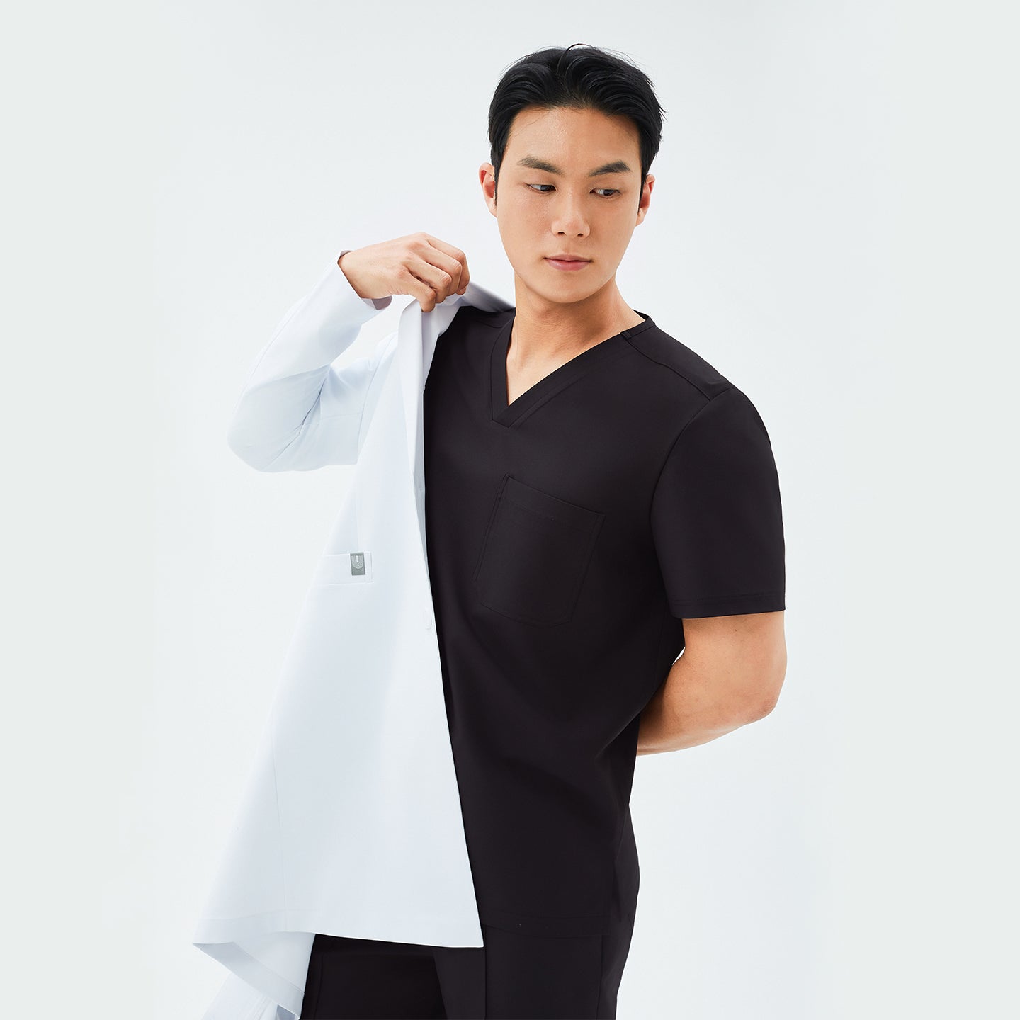 Model in a black scrub top, partially removing or putting on the TUL LCM-11 Men’s Long Lab Coat in White, revealing the coat’s interior label and smooth lining.