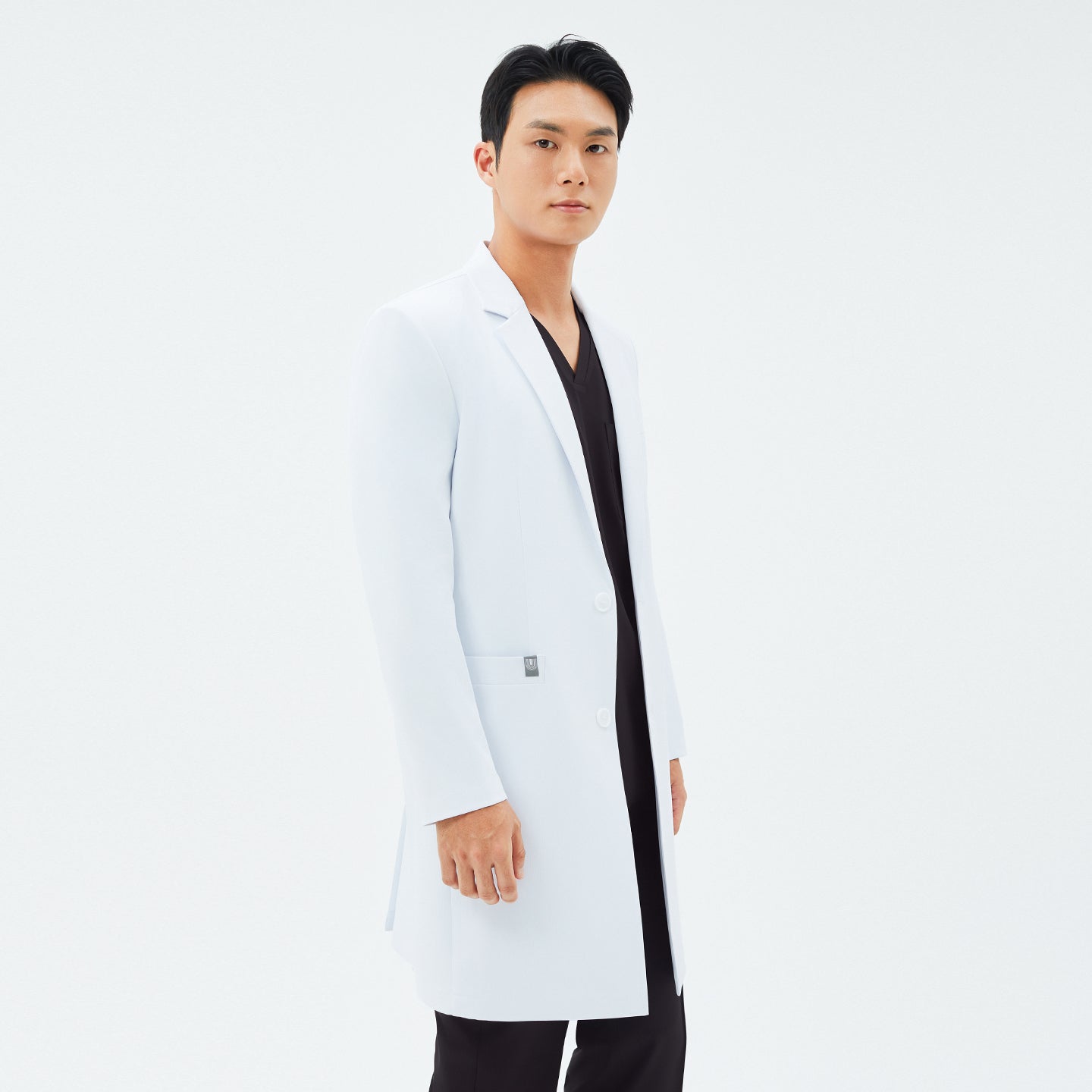 Image of Side angle of the TUL LCM-11 Men’s Long Lab Coat in White, showing a notched lapel, button closure, and fitted sleeves. The model wears black scrubs underneath.