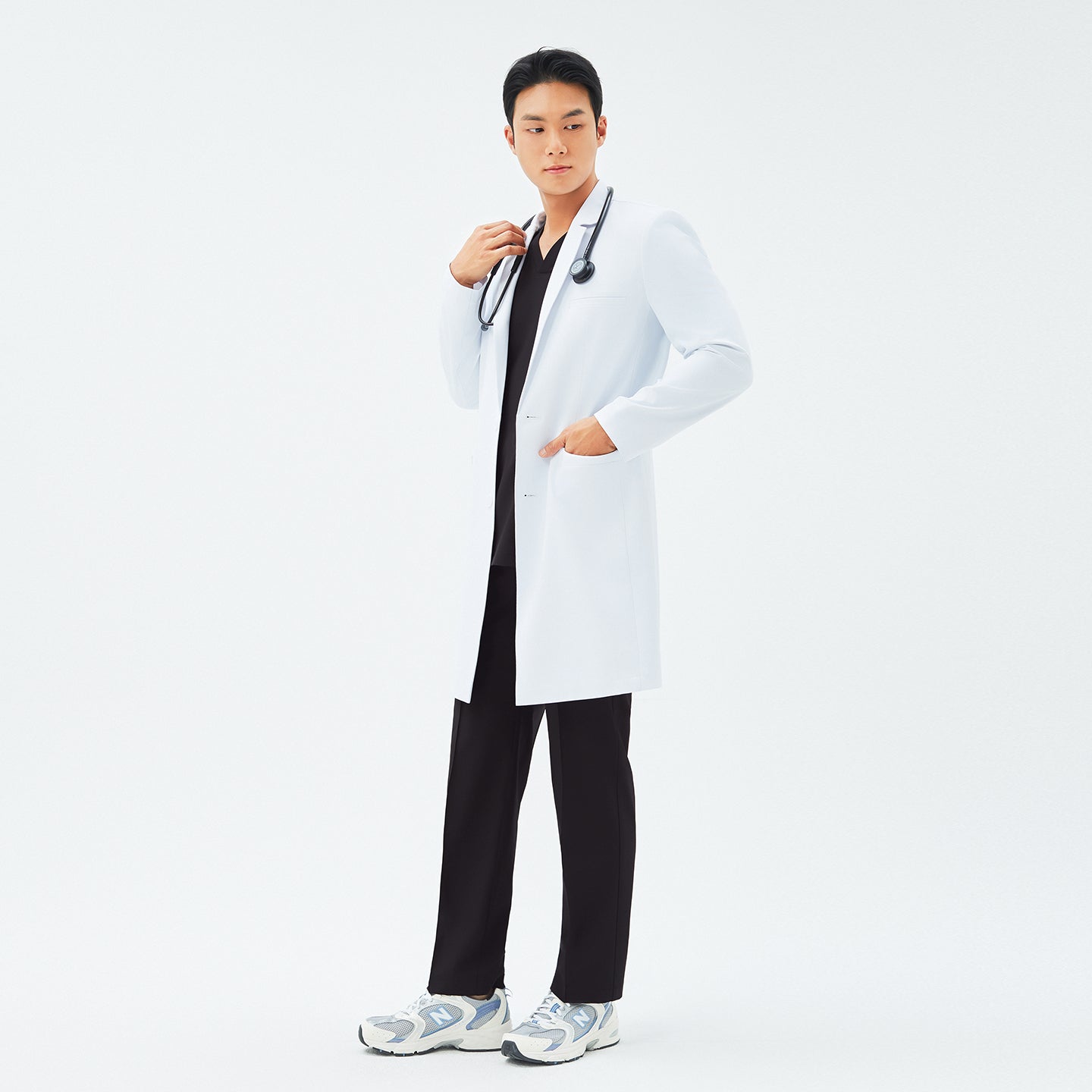 Male model wearing TUL LCM-11 Men’s Long Lab Coat in White over a black scrub set, standing with one hand in his coat pocket and a stethoscope around his neck.
