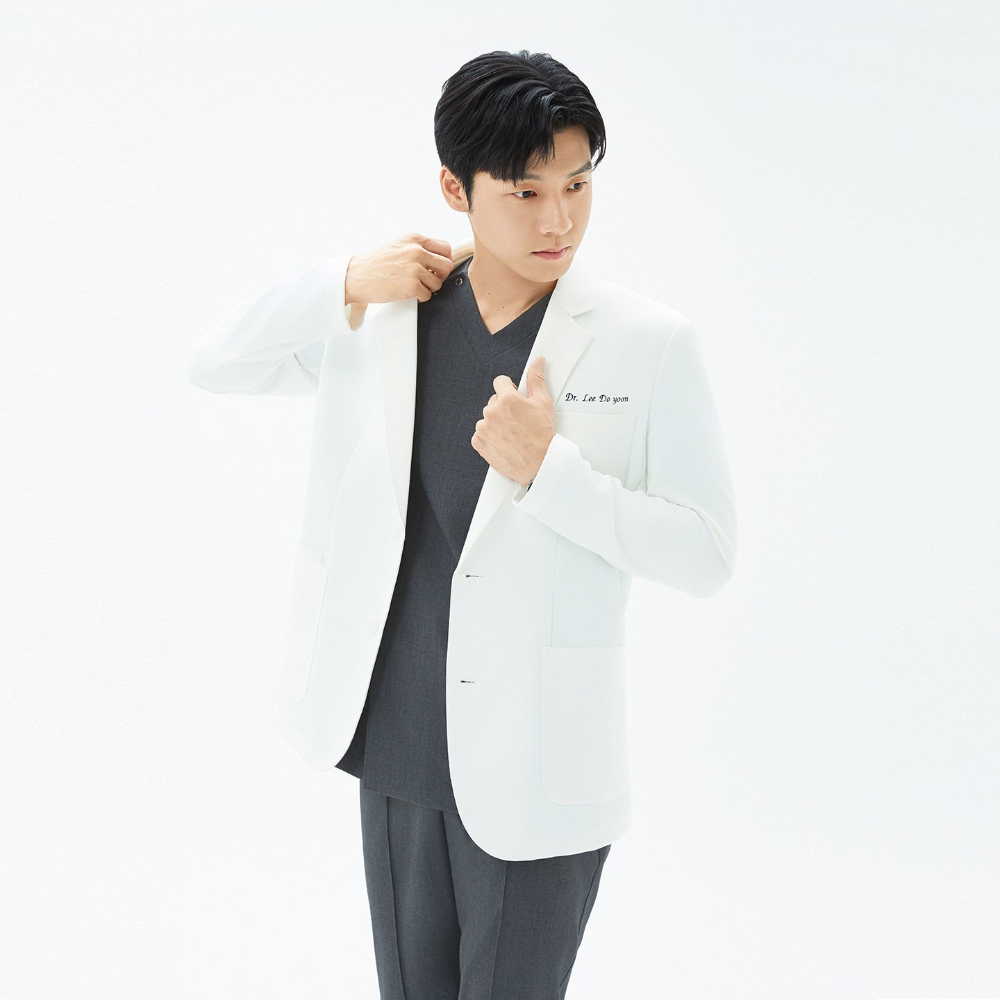 Image of Male model in a white ivory short lab coat adjusting the notched lapel collar, standing against a plain white background.
