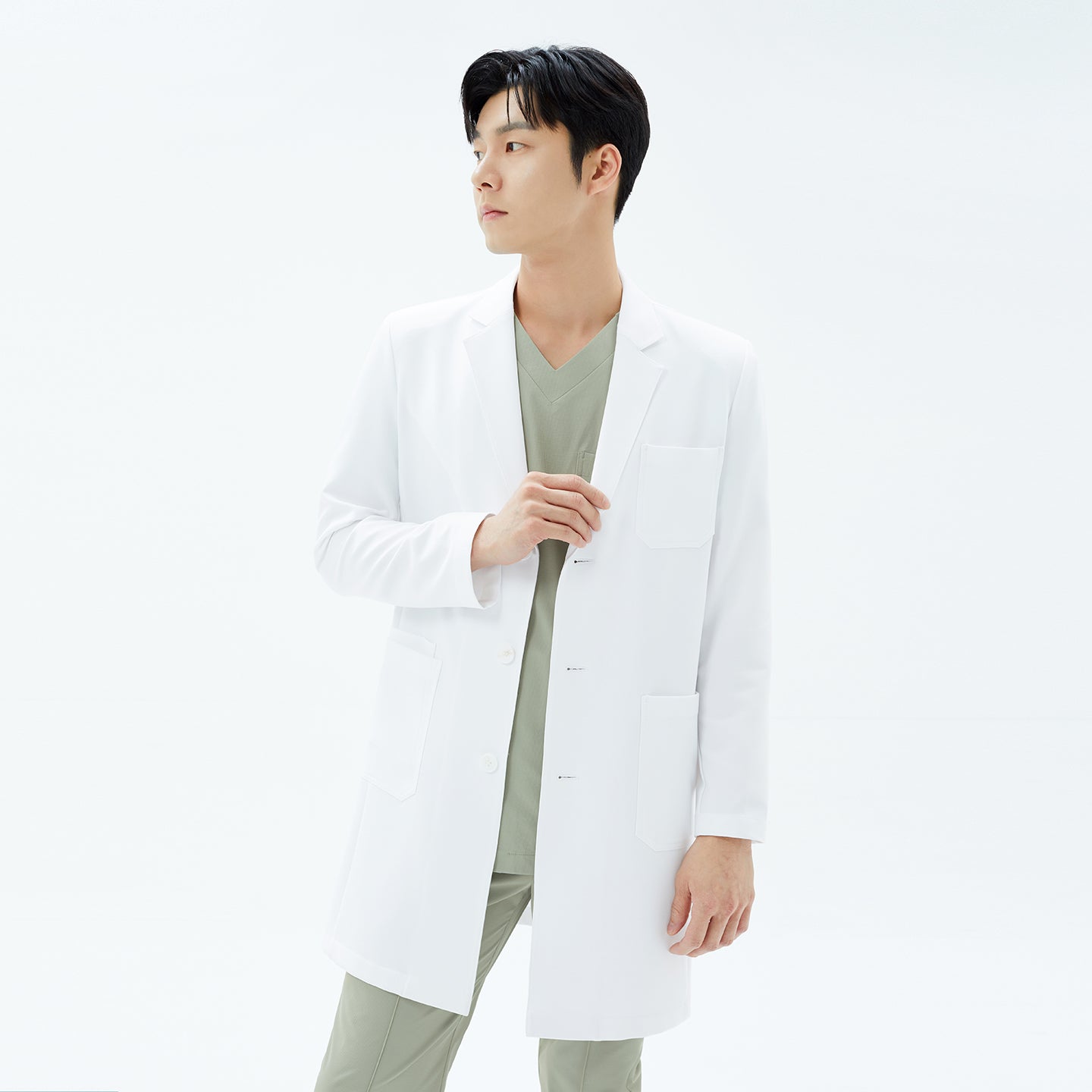 Image of A male model in a bright studio adjusts the lapel of his TUL Men’s LCM-15 Core Long Lab Coat in White, revealing a glimpse of sand khaki scrubs underneath.