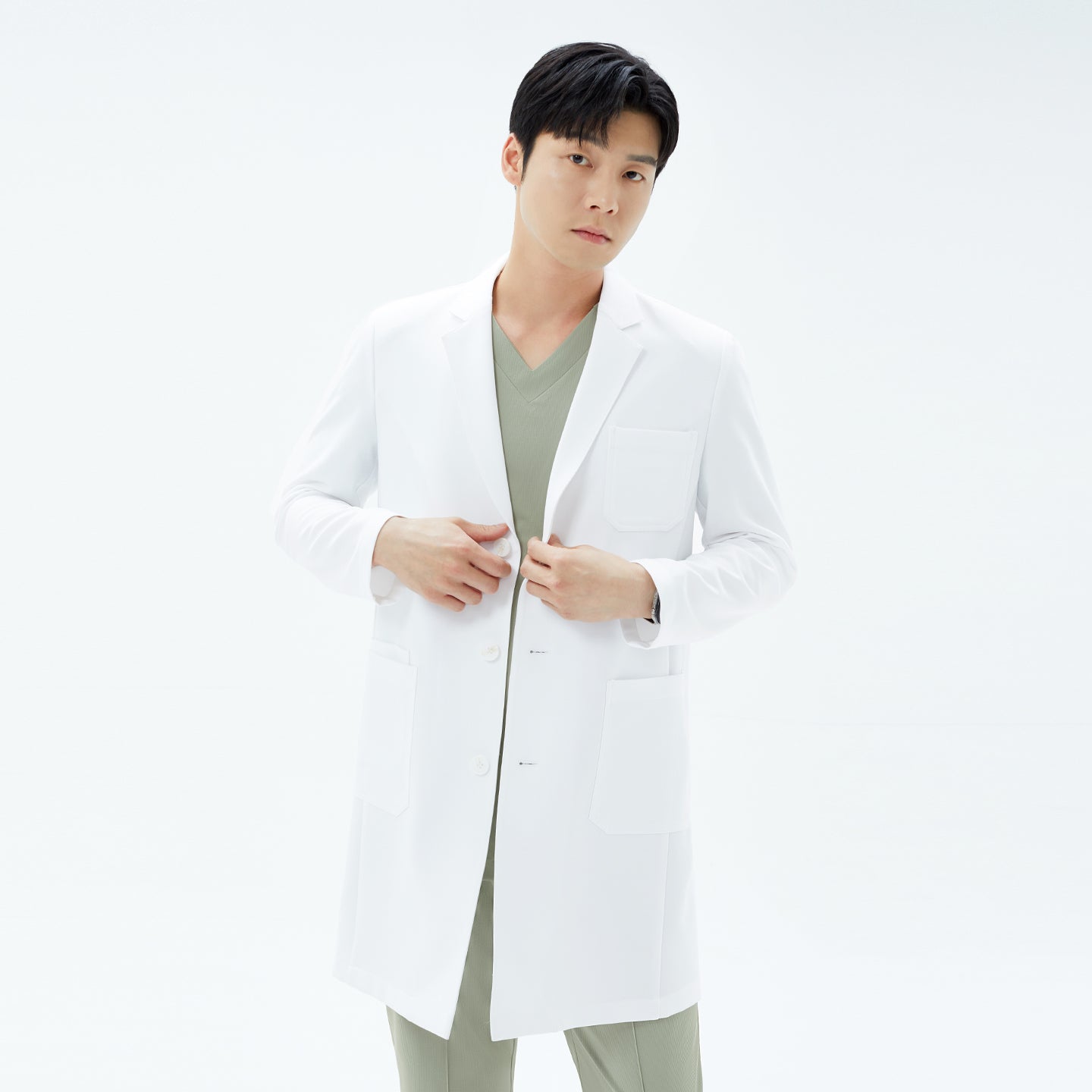 Image of Frontal shot of the model buttoning his white TUL Men’s LCM-15 Core Long Lab Coat over a khaki scrub top, gazing directly ahead.