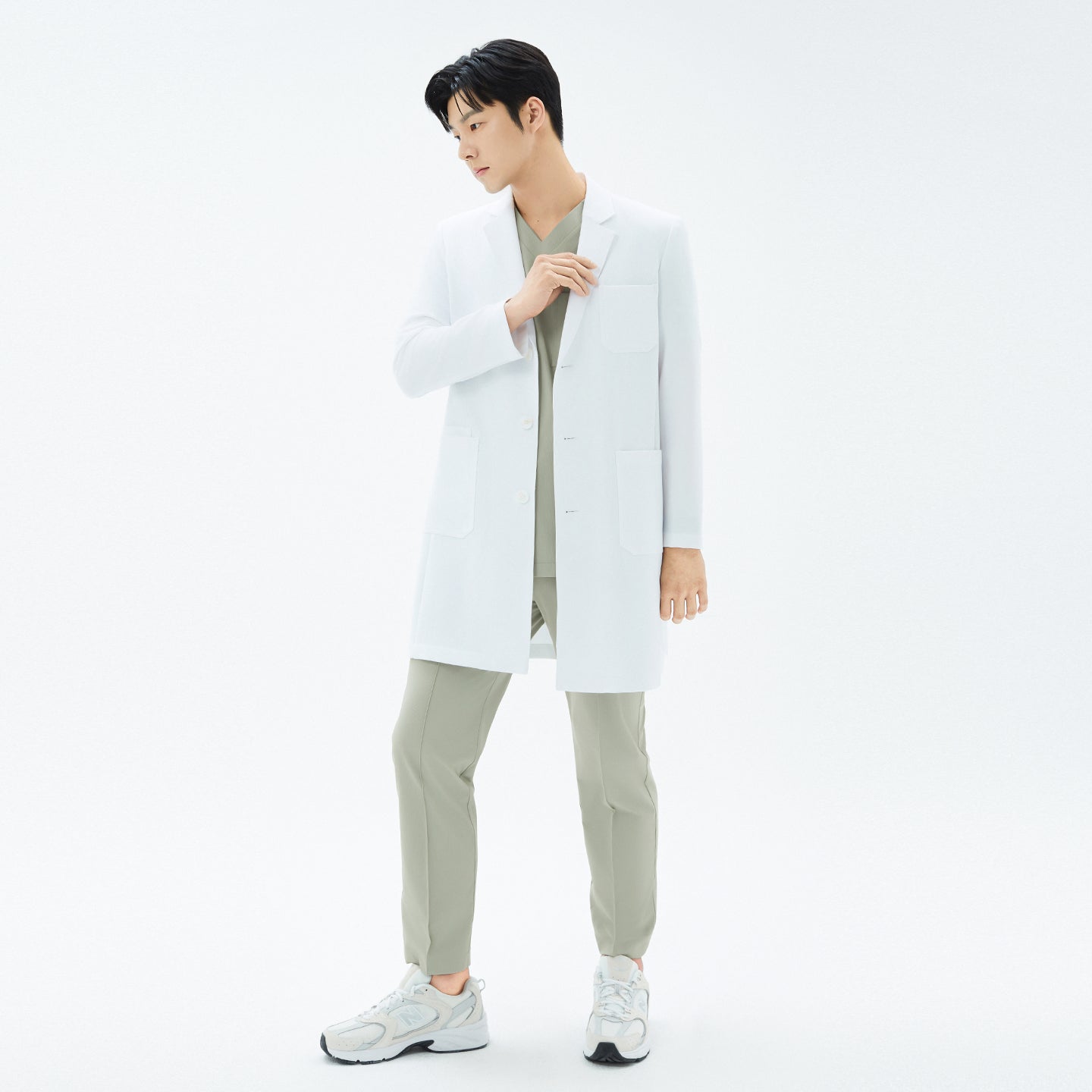 Image of Full-body image of the model in the white TUL Men’s LCM-15 Core Long Lab Coat and khaki scrubs, hand lifting the chest pocket, feet slightly apart.