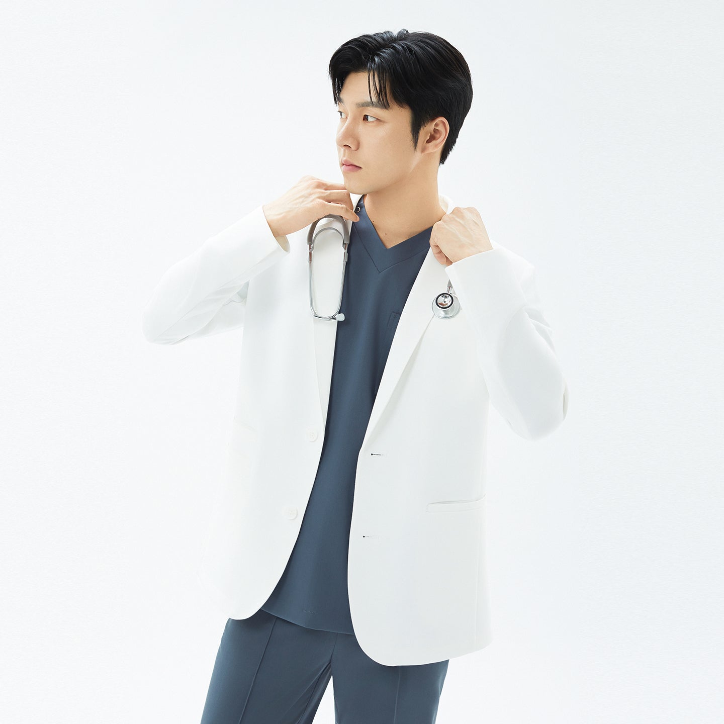 Image of Three-quarter view of the model draping a stethoscope around his neck while wearing the TUL Men’s LCM-17 UltraFit Short Lab Coat — White Ivory.