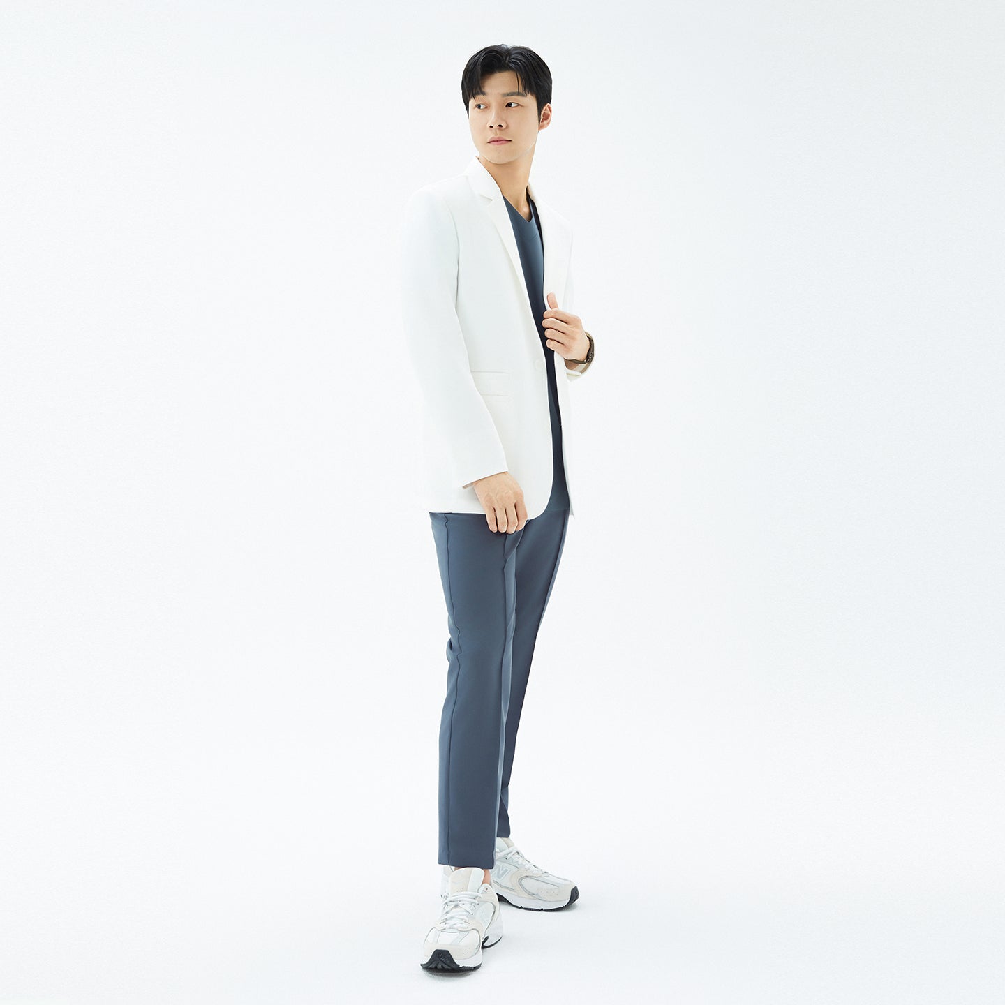 Image of Side full-length shot of a model standing with hands in pockets, wearing the TUL Men’s LCM-17 UltraFit Short Lab Coat in White Ivory over scrub pants.